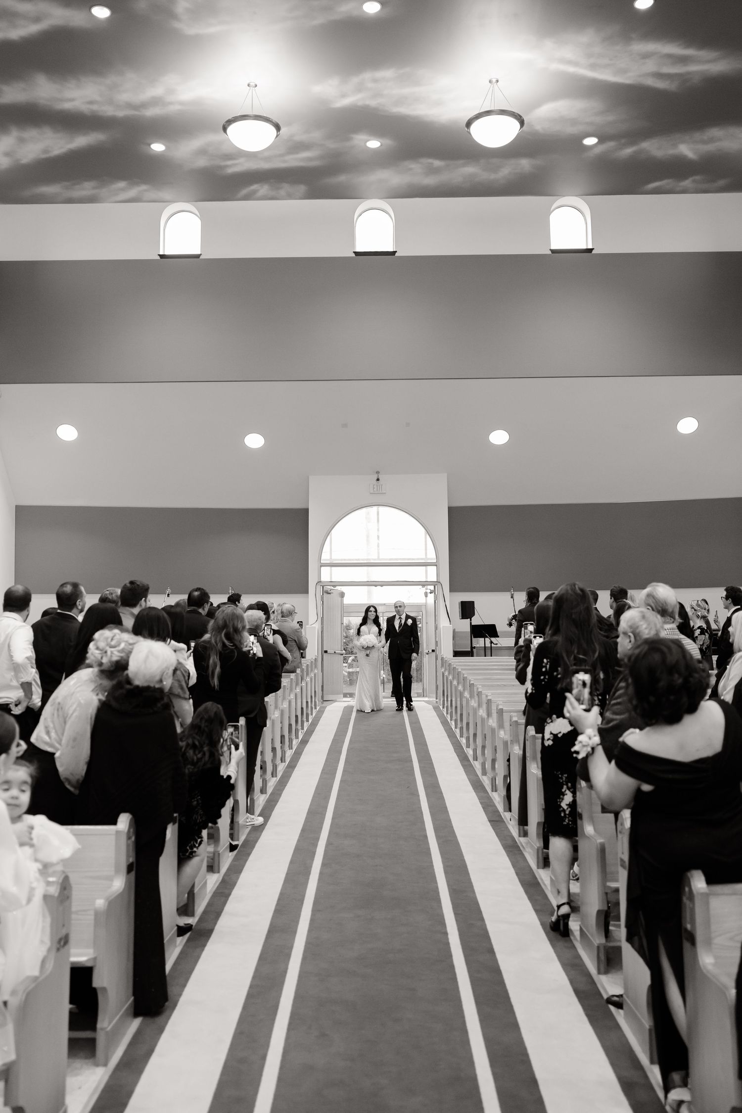 Black and white photo of wedding ceremony processional in church with guests lining the aisle.