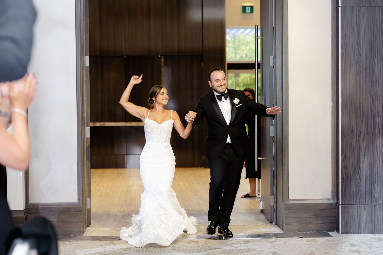 A bride and groom make their wedding entrance through wooden doors with joyful expressions and celebratory gestures.
