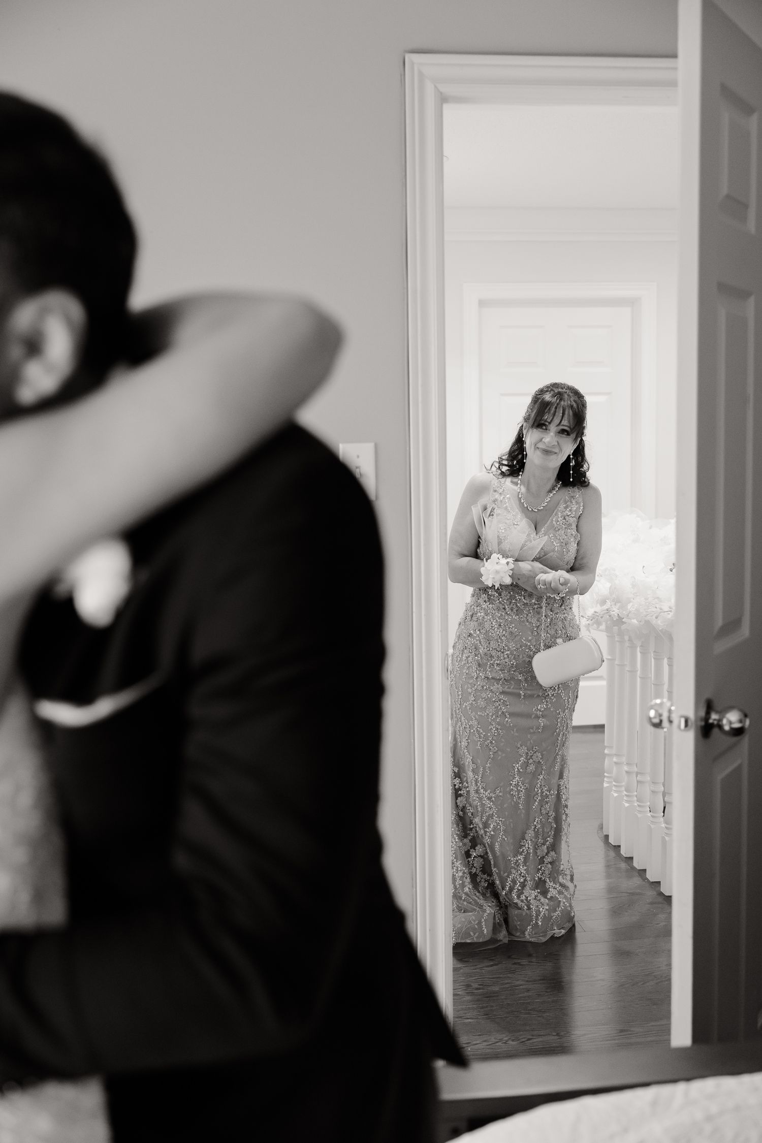 Black and white photo of someone in a formal gown seen through a doorway reflection in a mirror.