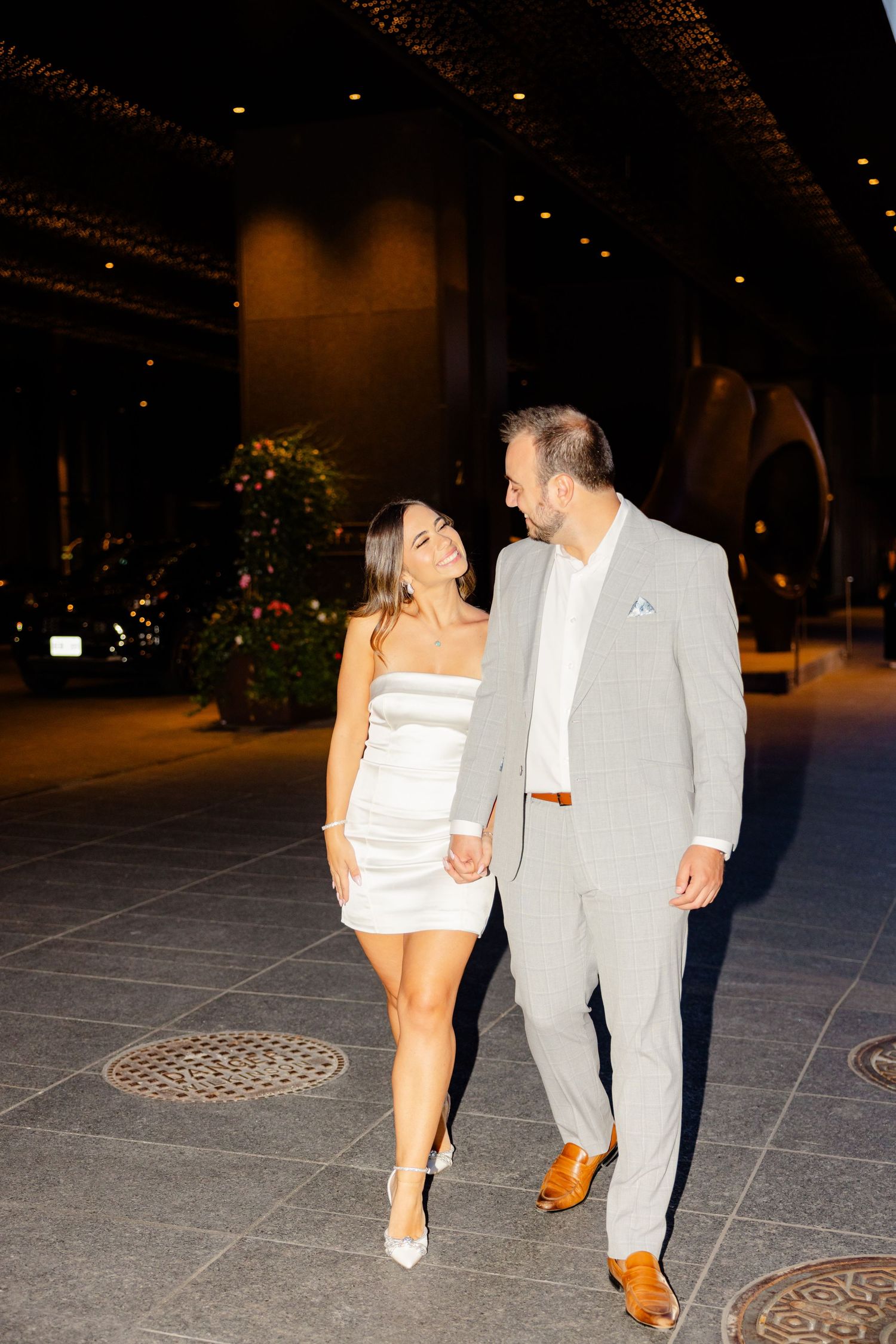 Couple in evening attire walking together, sharing a laugh in an elegantly lit indoor venue.