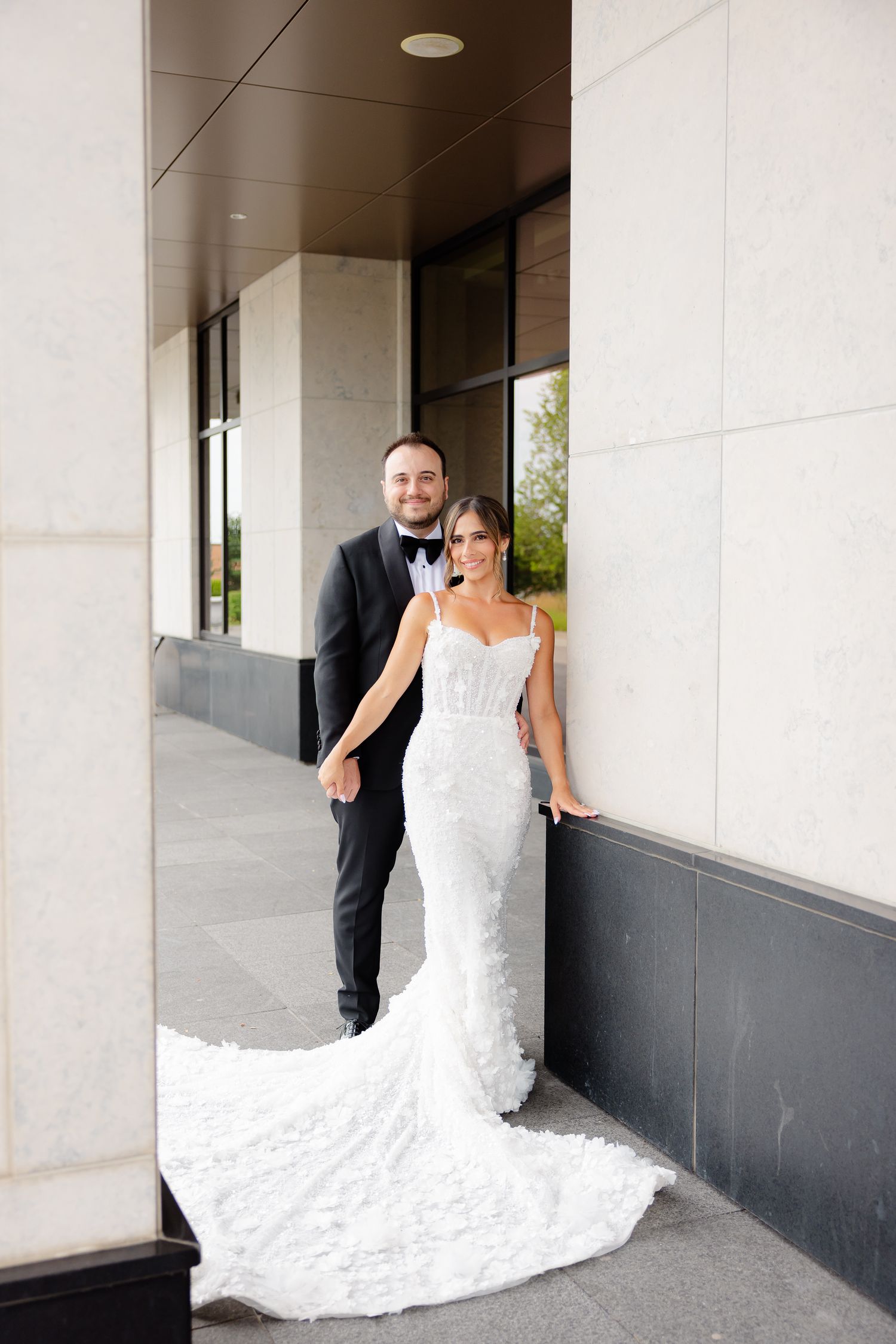 A bride in a fitted lace wedding gown with long train poses with groom in black suit outside a modern building.