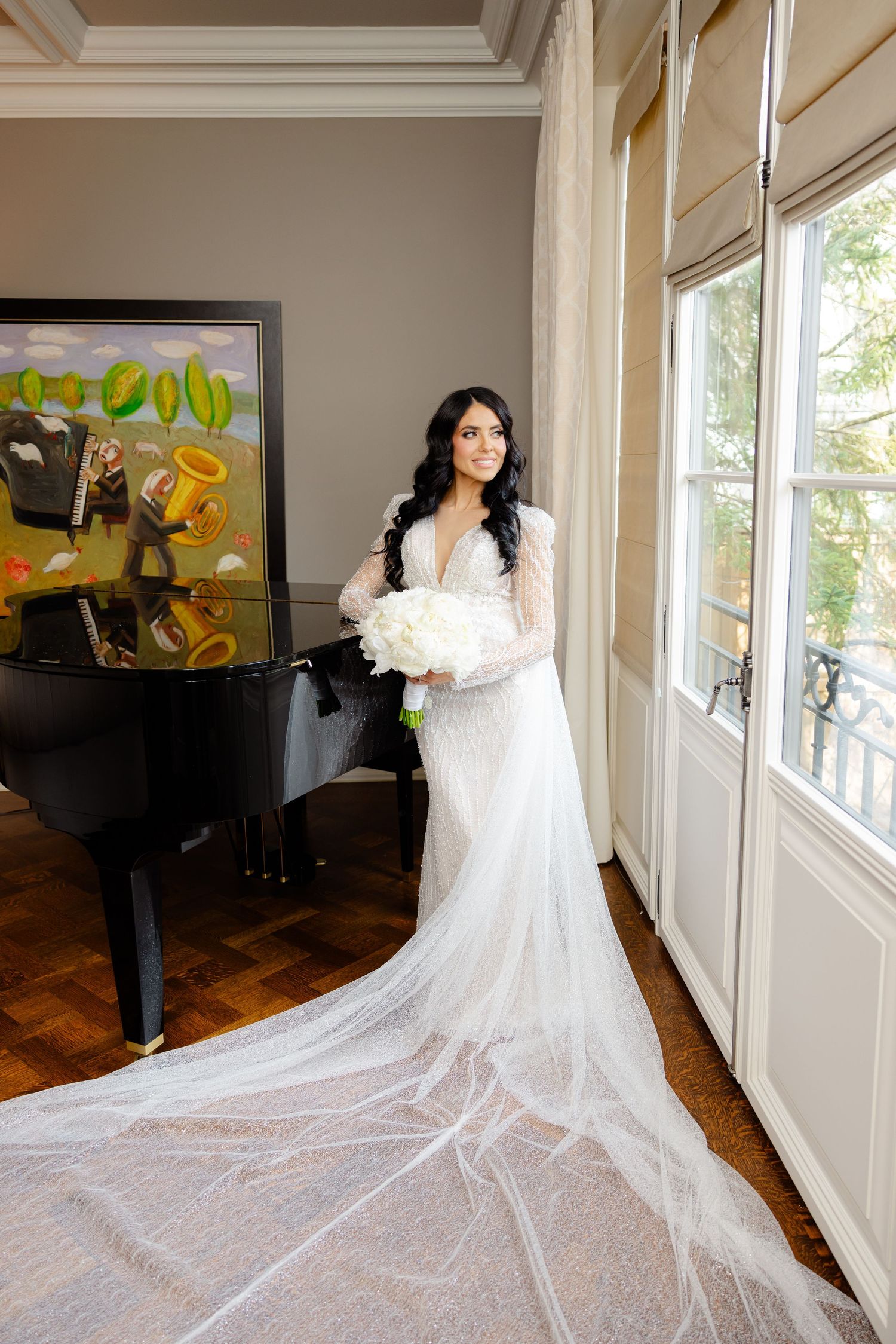 Bride in lace gown holding white bouquet poses by piano and French doors.