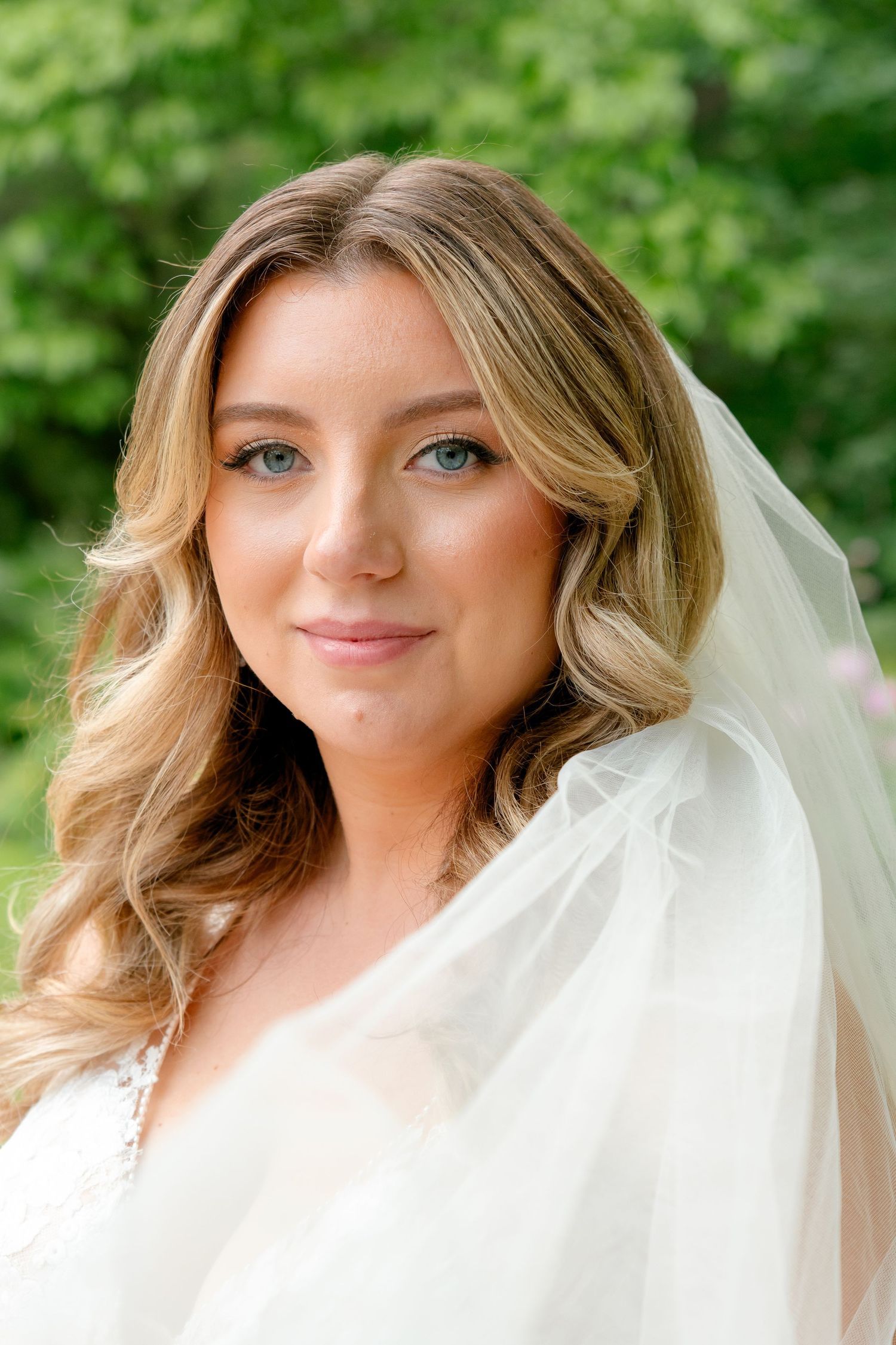 A bride wearing a flowing white veil smiles at the camera against a leafy green background.
