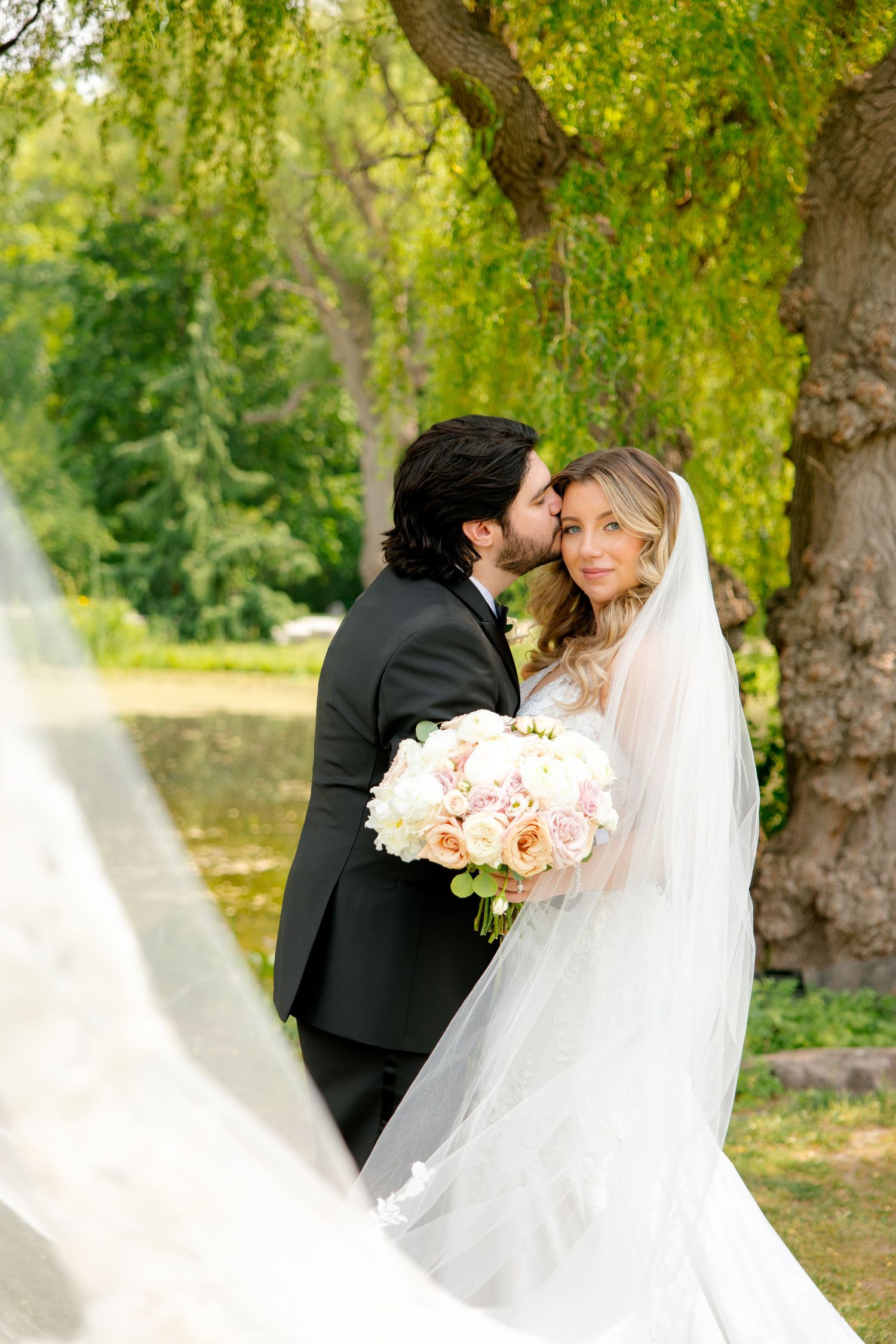 Romantic garden wedding photo with flowing veil and soft pink roses under willow branches.