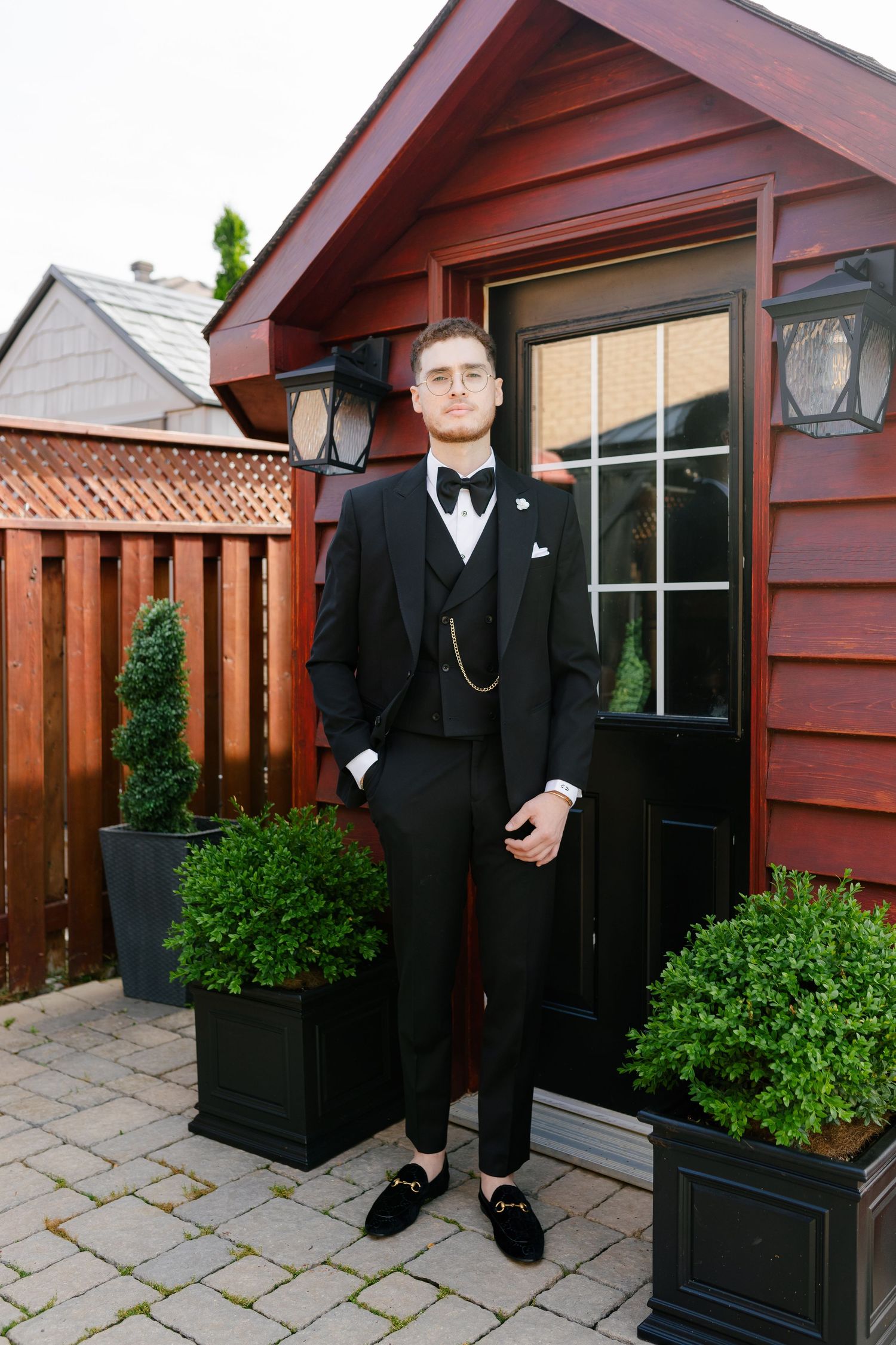 A well-dressed person in a formal black tuxedo stands in front of a red wooden shed with potted plants.