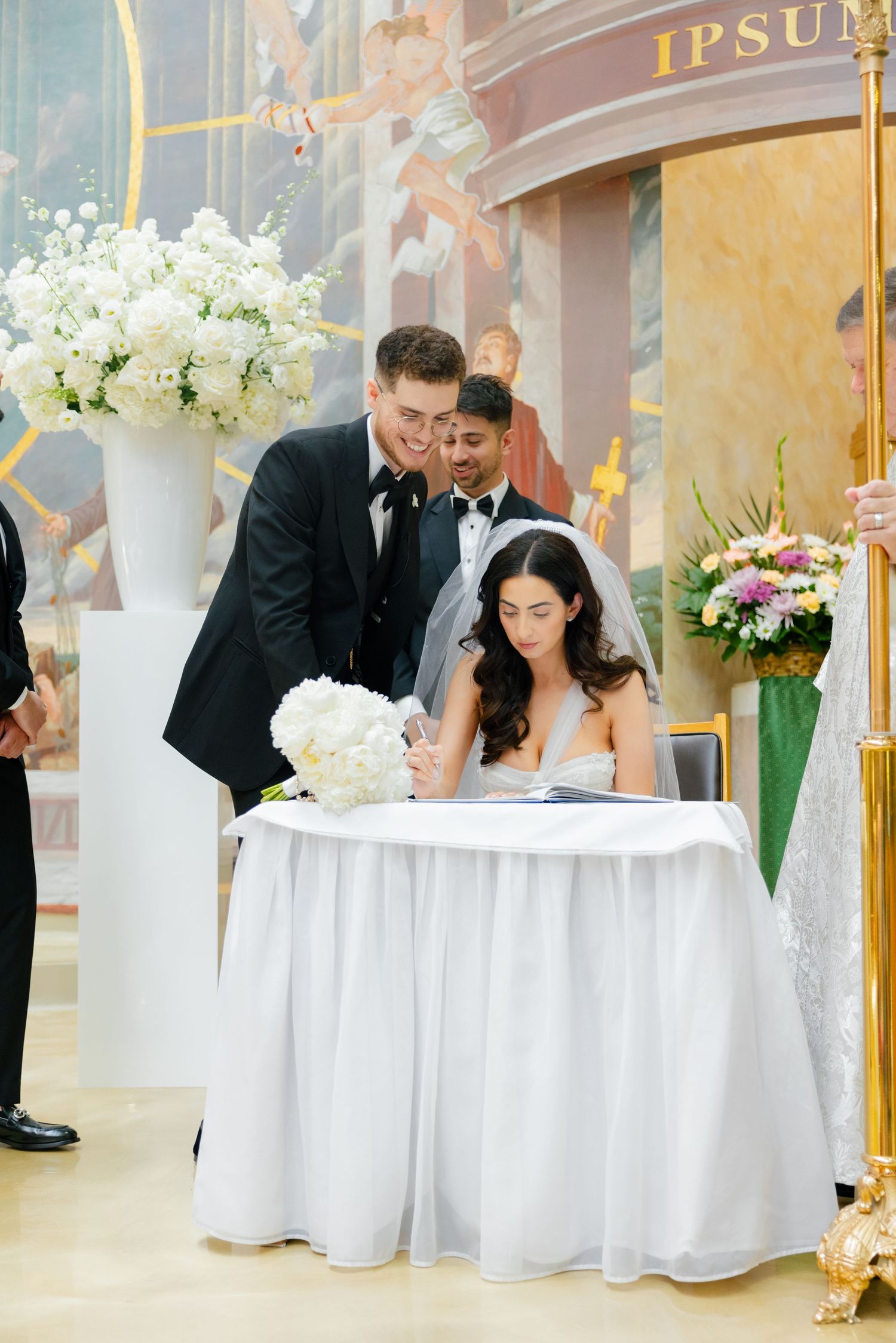 A wedding ceremony moment captured during document signing with floral arrangements.