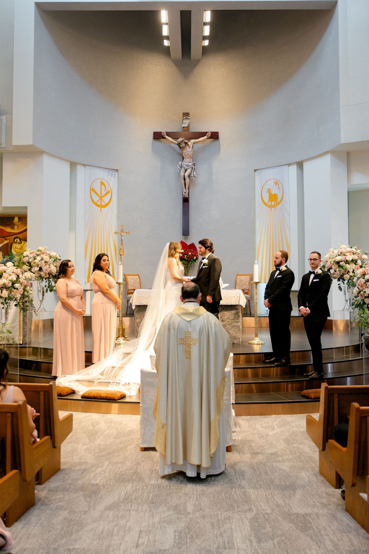 Wedding ceremony in a modern church with crucifix, floral arrangements, and wedding party on elevated altar.