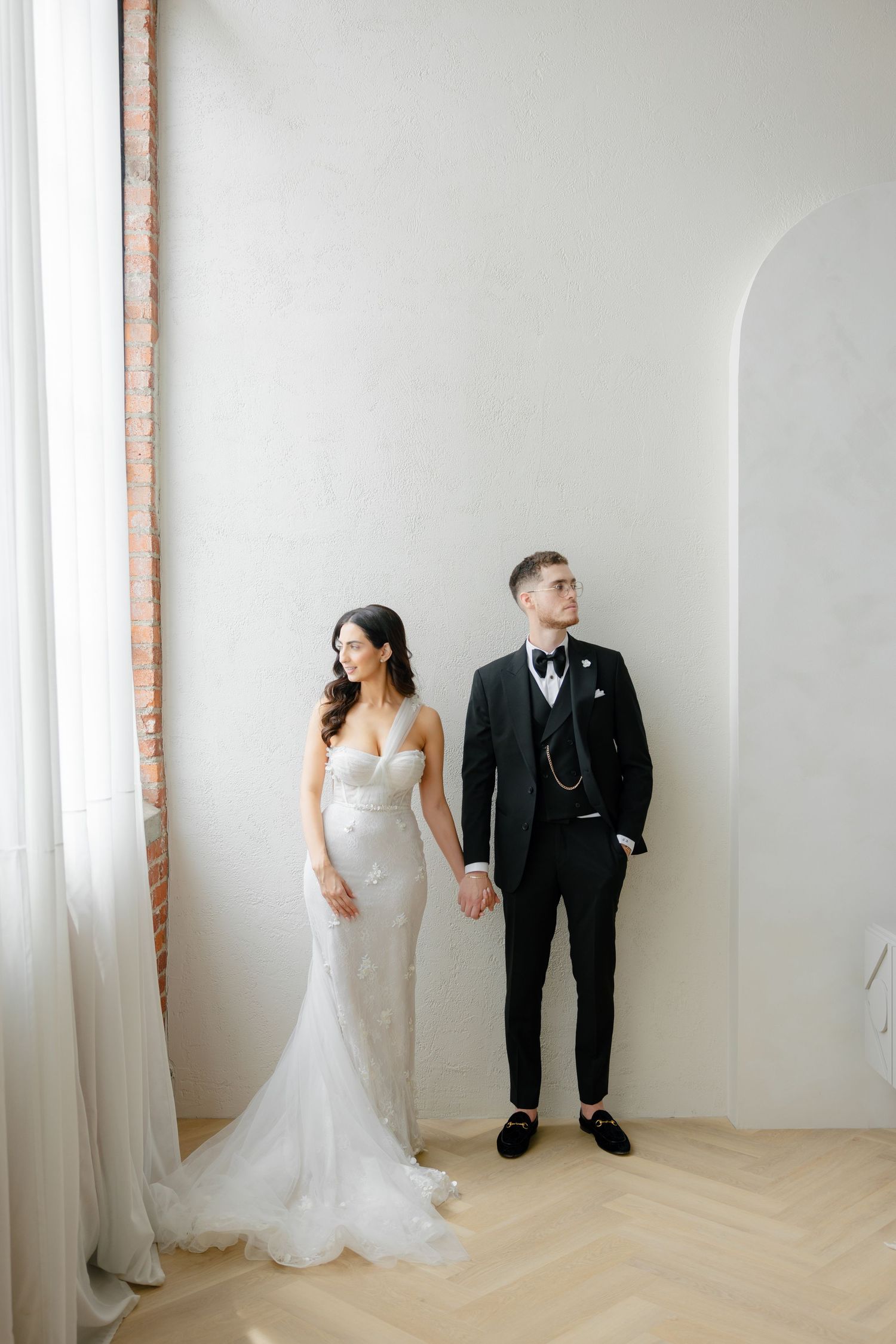 Modern wedding couple poses against white wall in elegant attire.