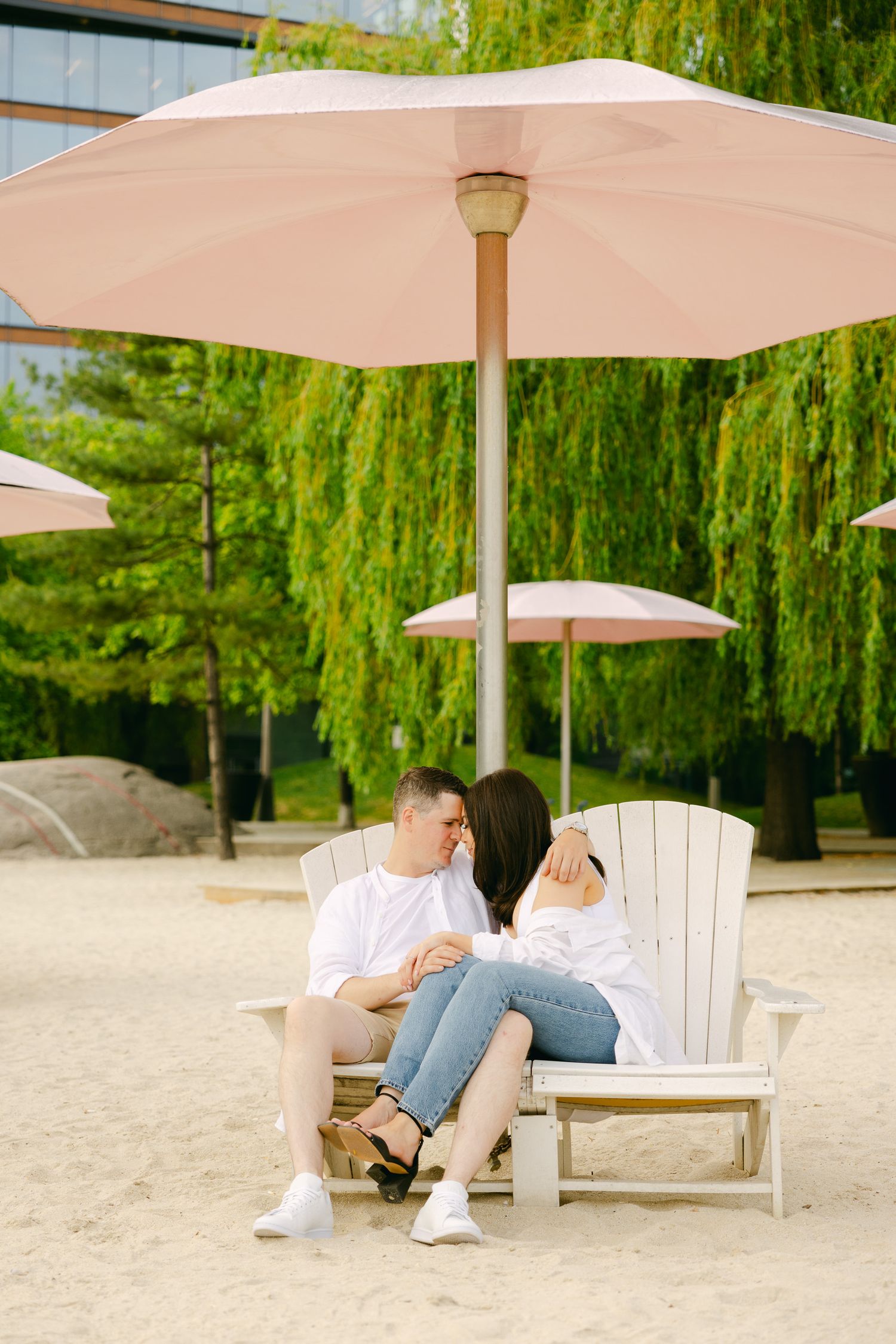 A couple relaxes on an Adirondack chair under a pink umbrella near weeping willow trees.