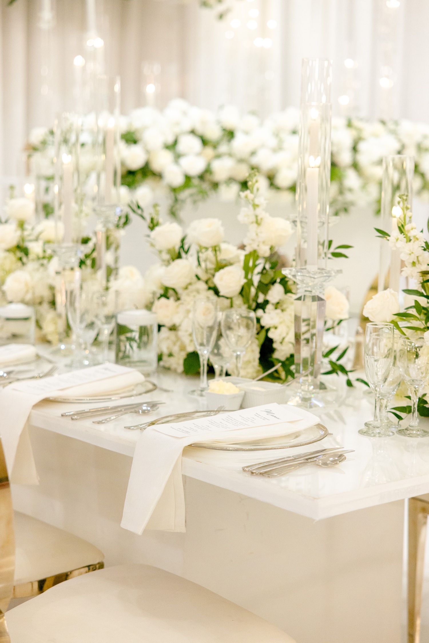 Sophisticated white reception table with crystal candleholders and lush floral arrangements.