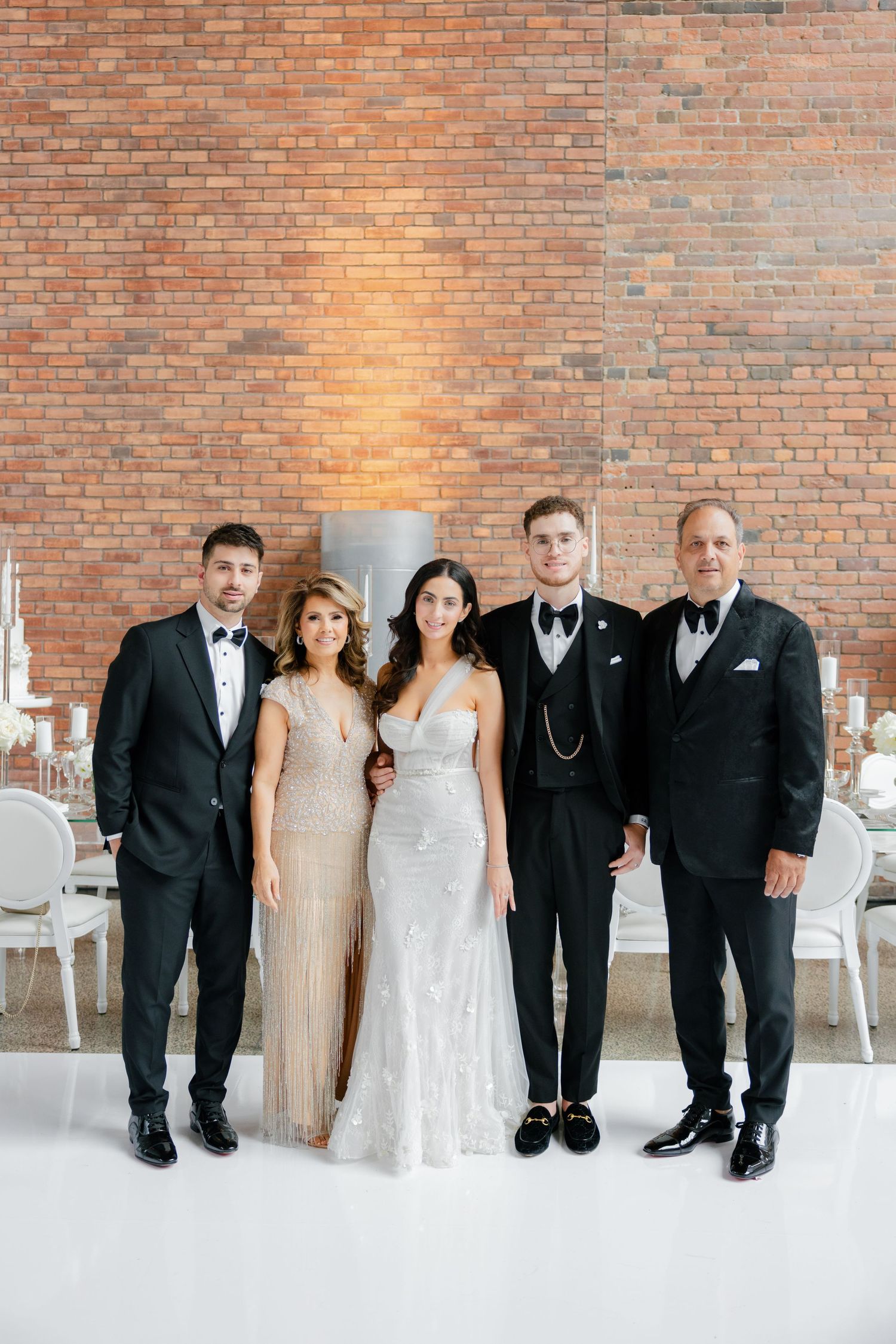 Wedding party in formal attire poses together with elegant white chairs and brick wall background.