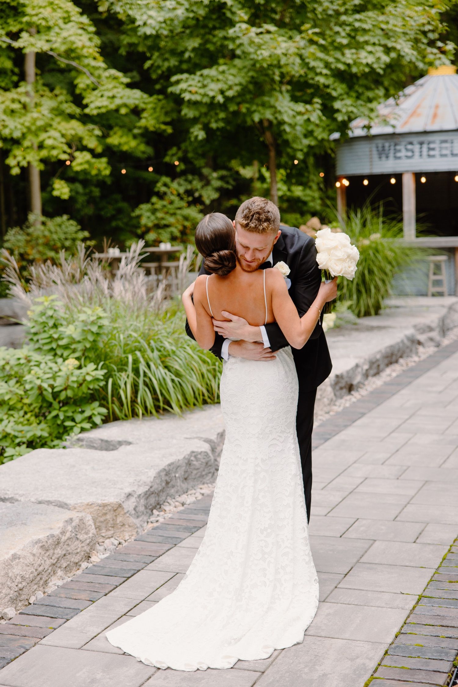 Couple in wedding attire embraces on garden path with white bouquet and rustic venue in background.