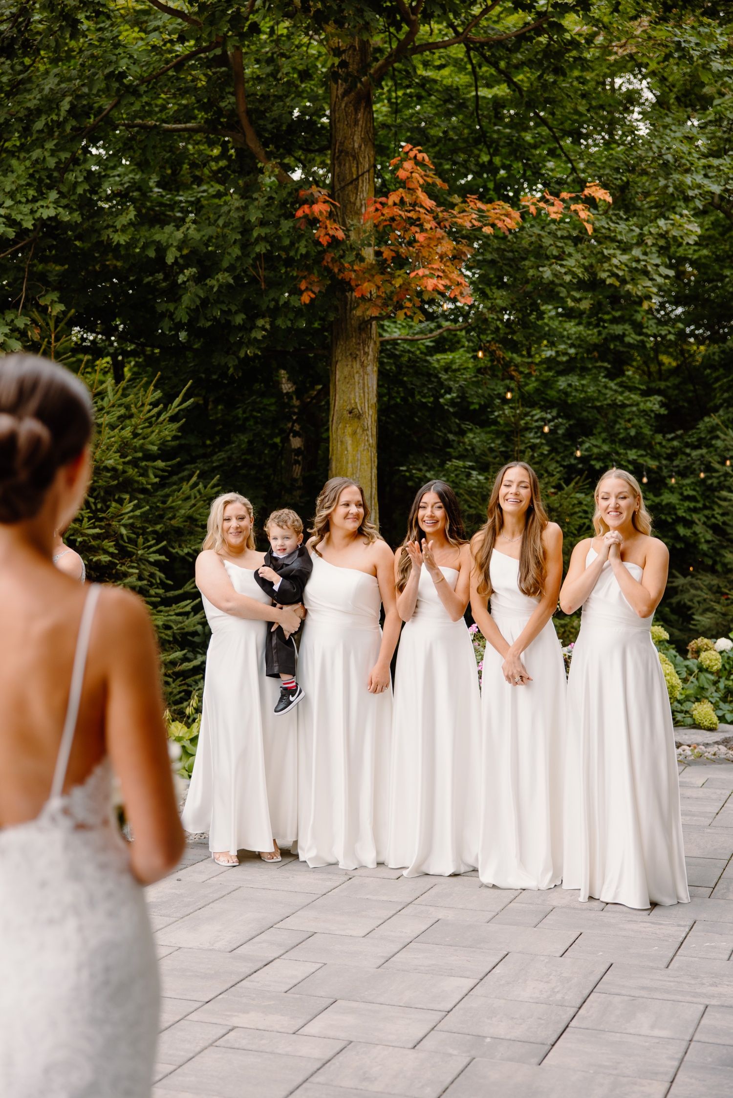 Group of bridesmaids in white dresses gathered on stone patio with autumn foliage in background.