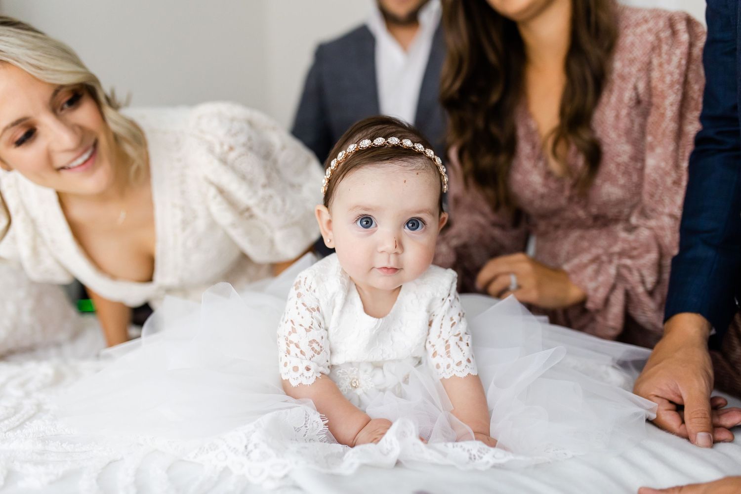Baby in white lace dress crawls on white bedding while adults look on in bedroom setting.