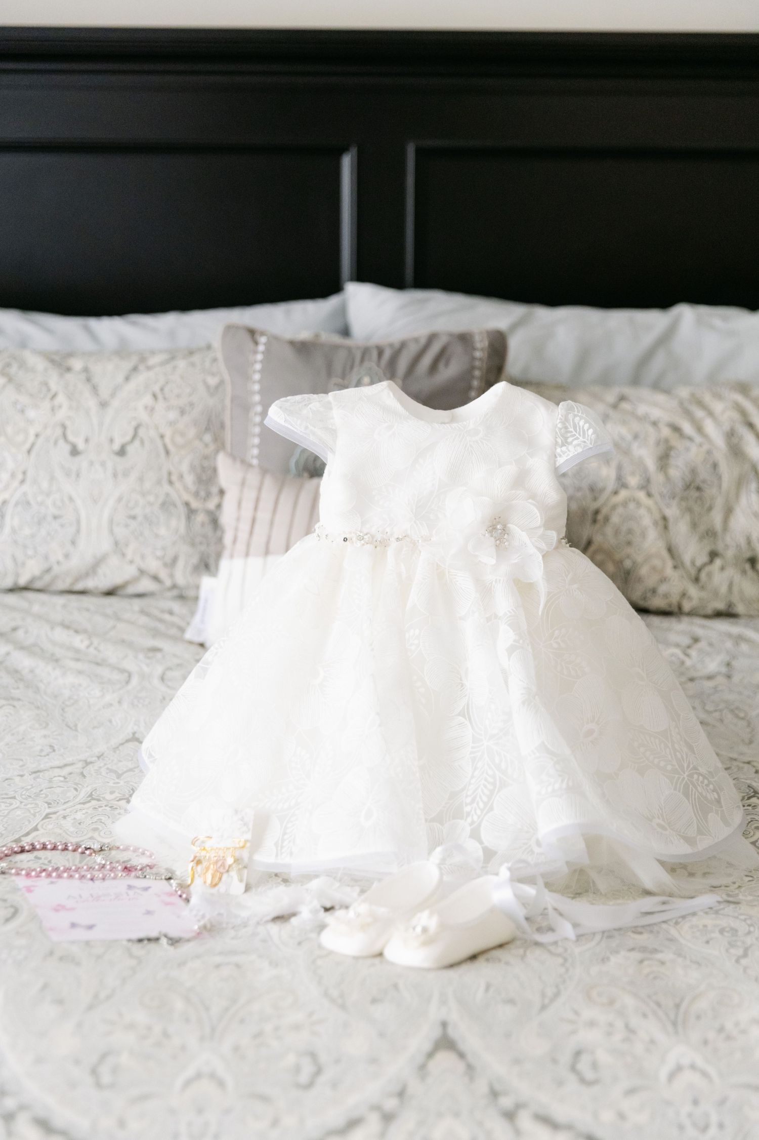 A delicate white baptismal dress and shoes laid out on a white textured bedspread.