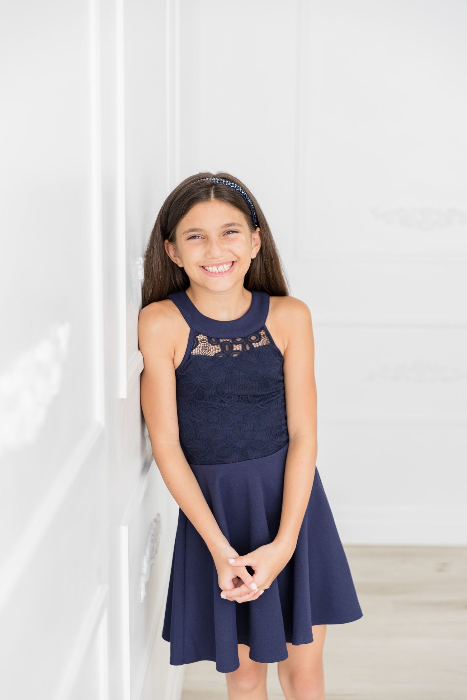 Girl in navy lace dress poses against white wall in natural light.