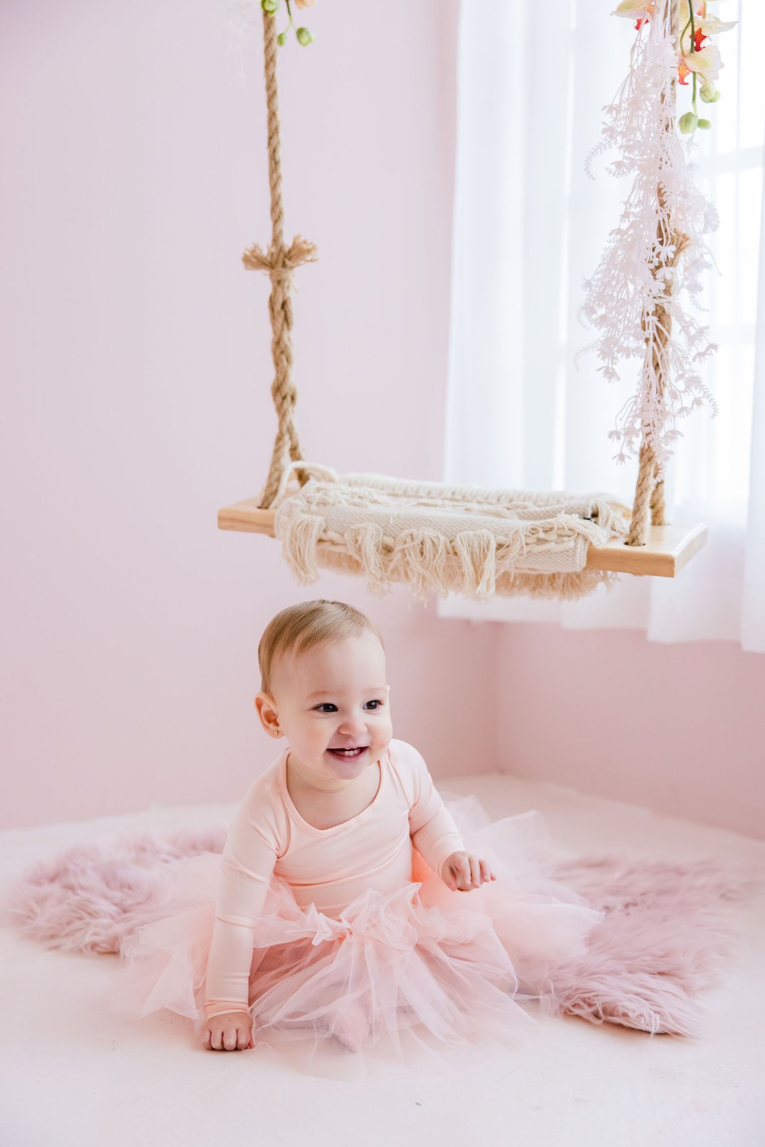 Toddler in pink tutu sits beneath decorative macrame swing in pink room.