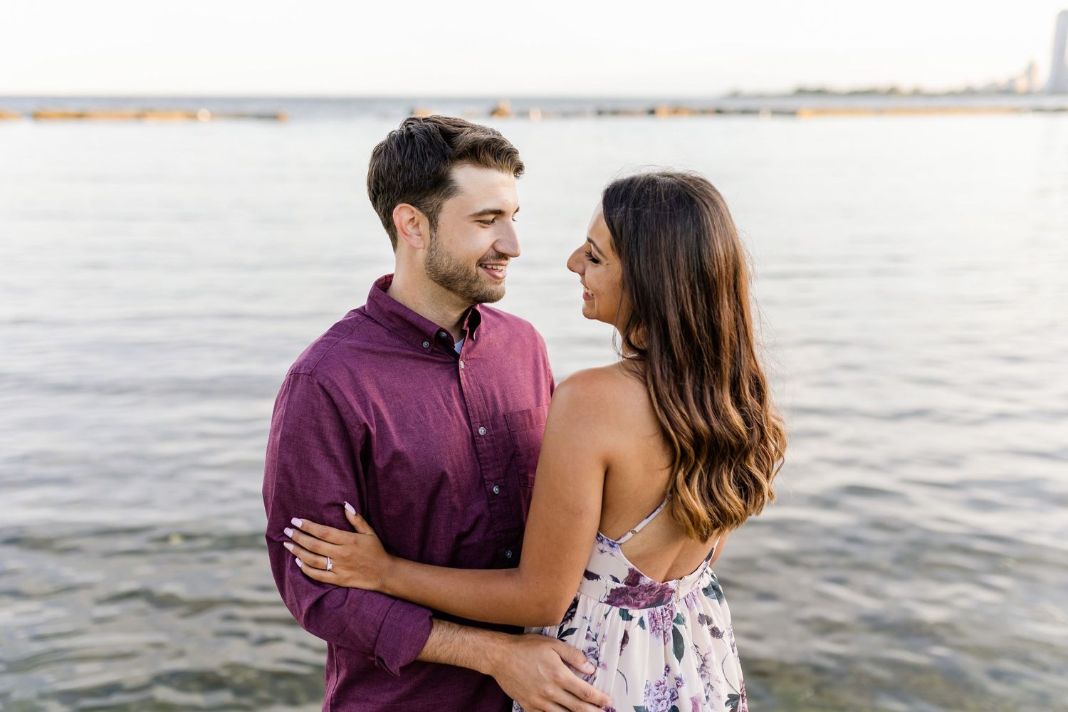 Romantic couple embracing by waterfront at sunset, wearing coordinated burgundy and floral outfits.
