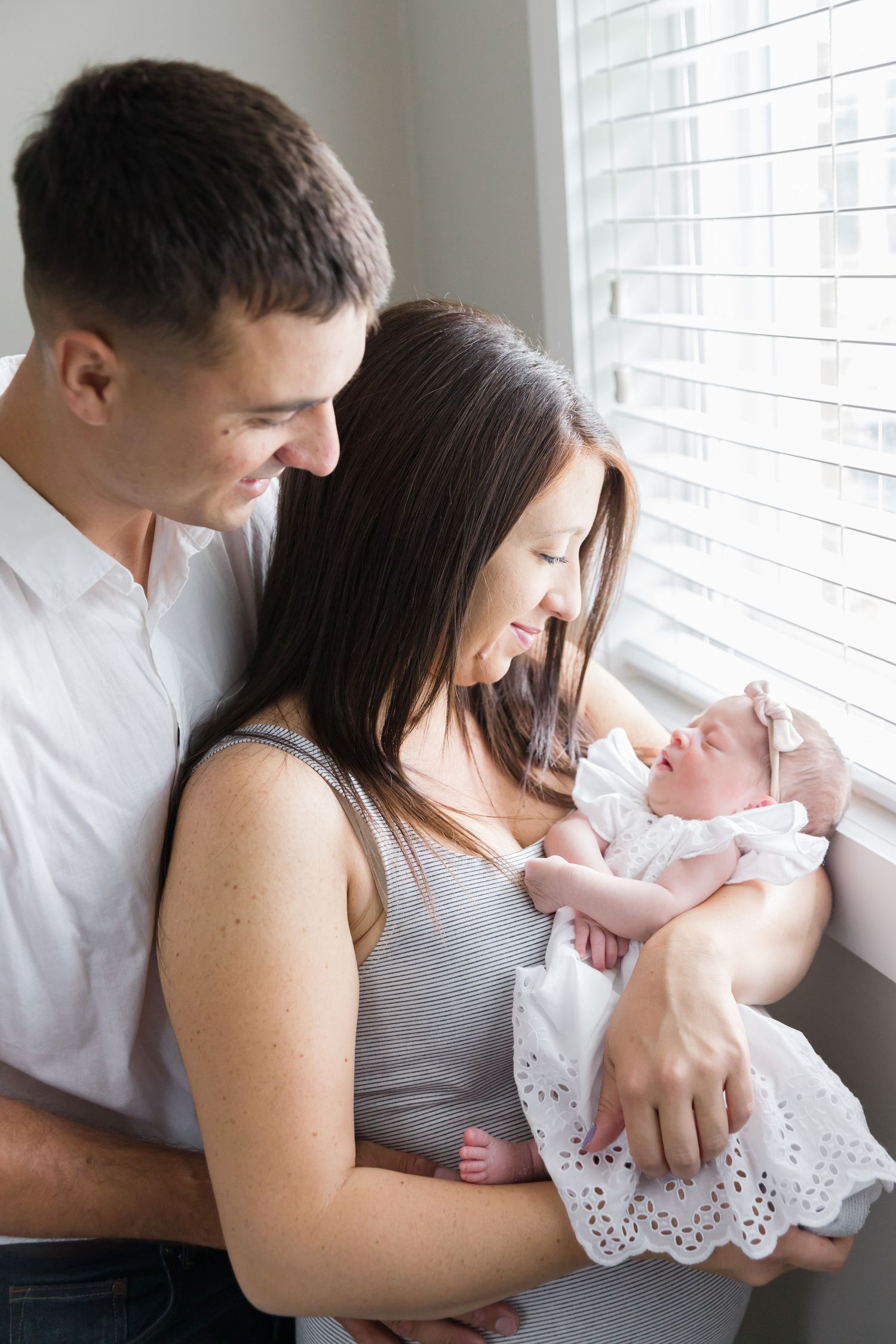 Parents sharing intimate moment with their newborn baby by window bathed in soft natural light.