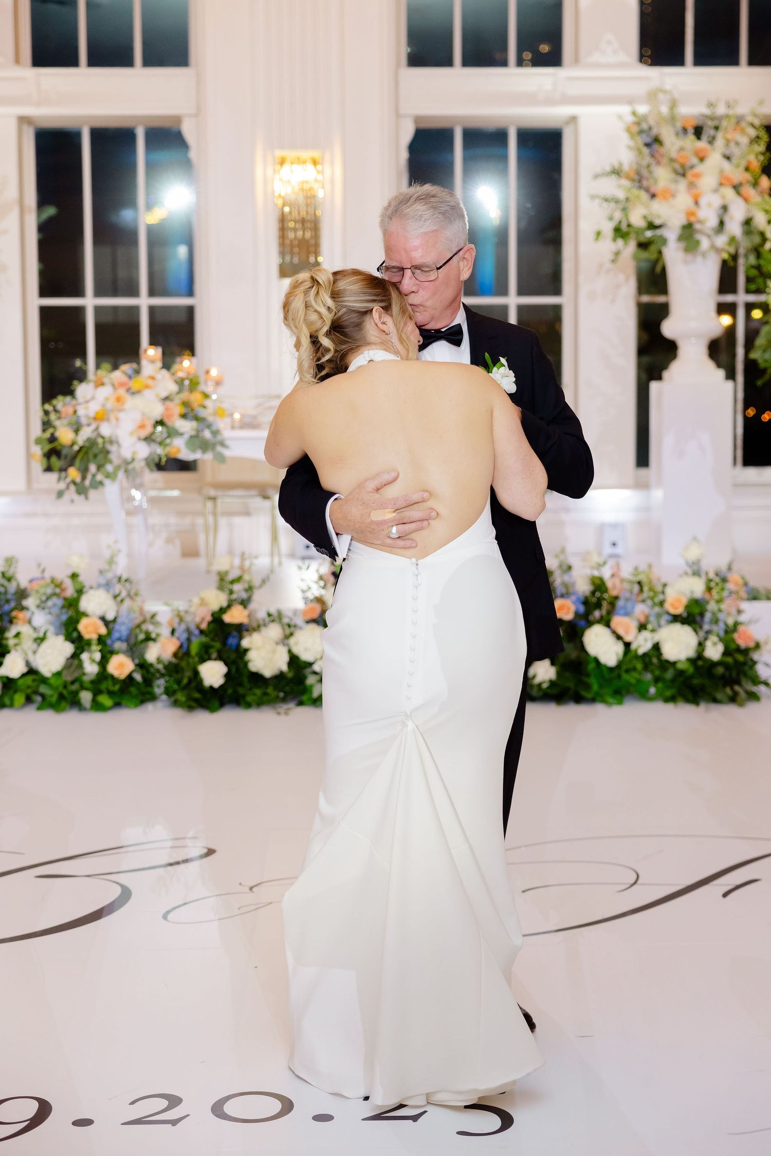 Couple dancing in an elegant white ballroom with floral arrangements and decorative floor design.