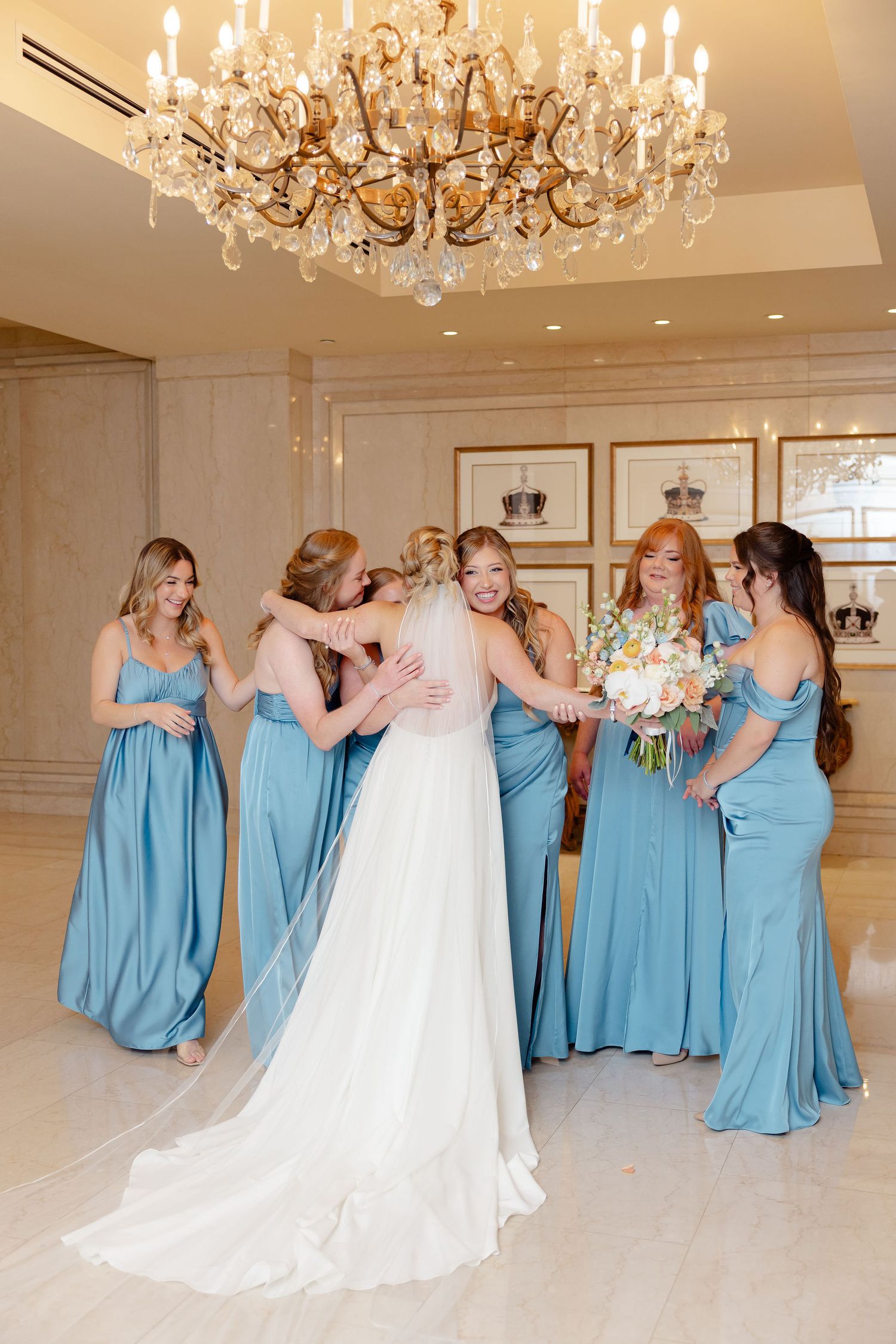 Bridesmaids in light blue dresses react joyfully to seeing the bride in her wedding gown under crystal chandeliers.
