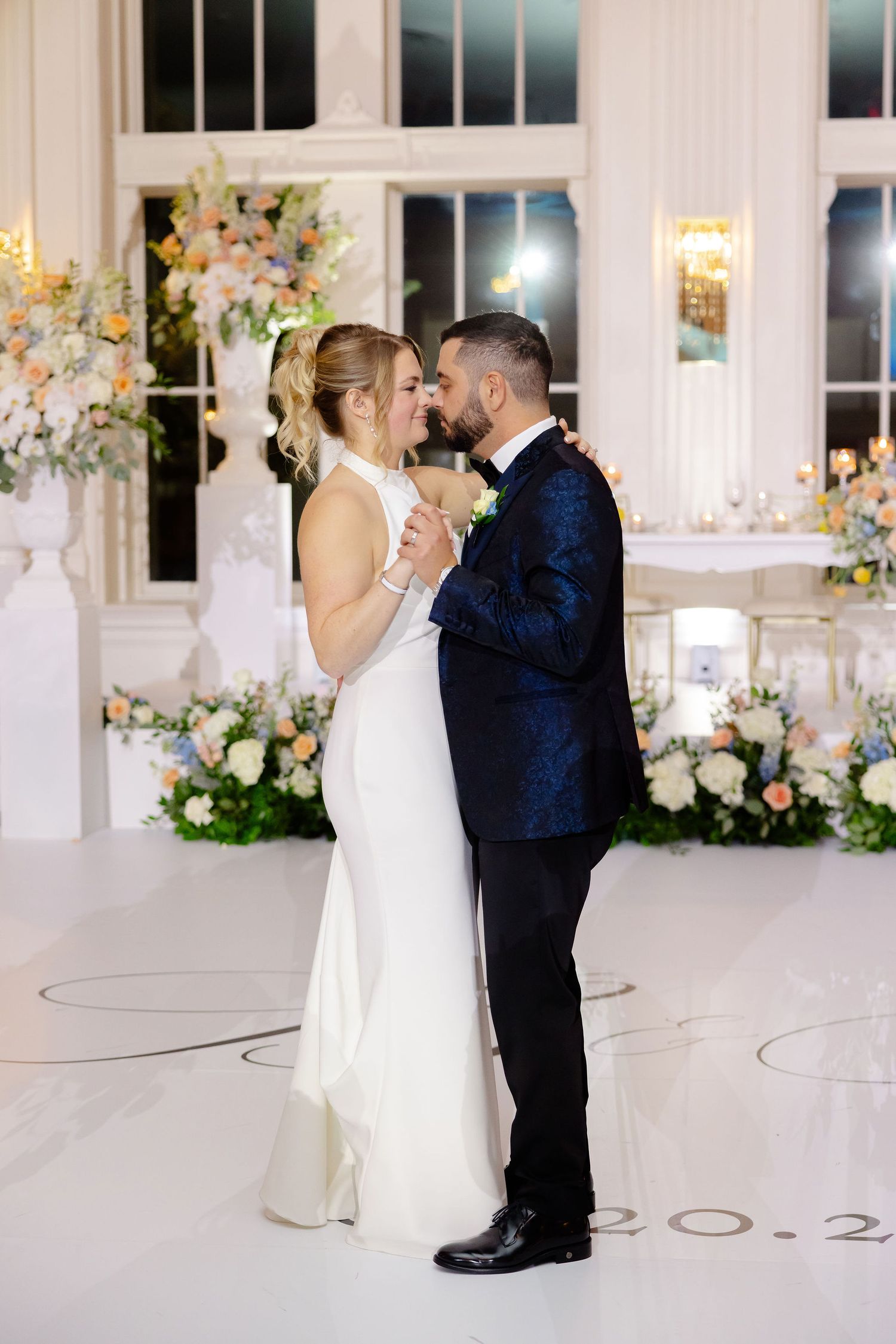 Wedding couple dancing in a bright ballroom with romantic floral decor and tall windows.
