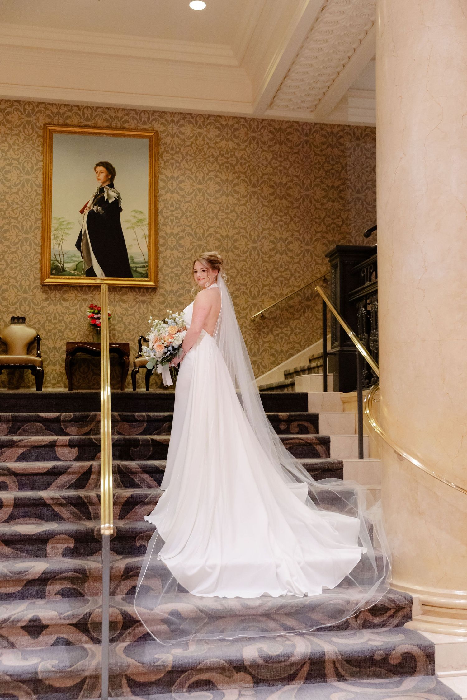 A flowing wedding dress train and veil create an elegant silhouette against a patterned carpet and wallpapered interior.