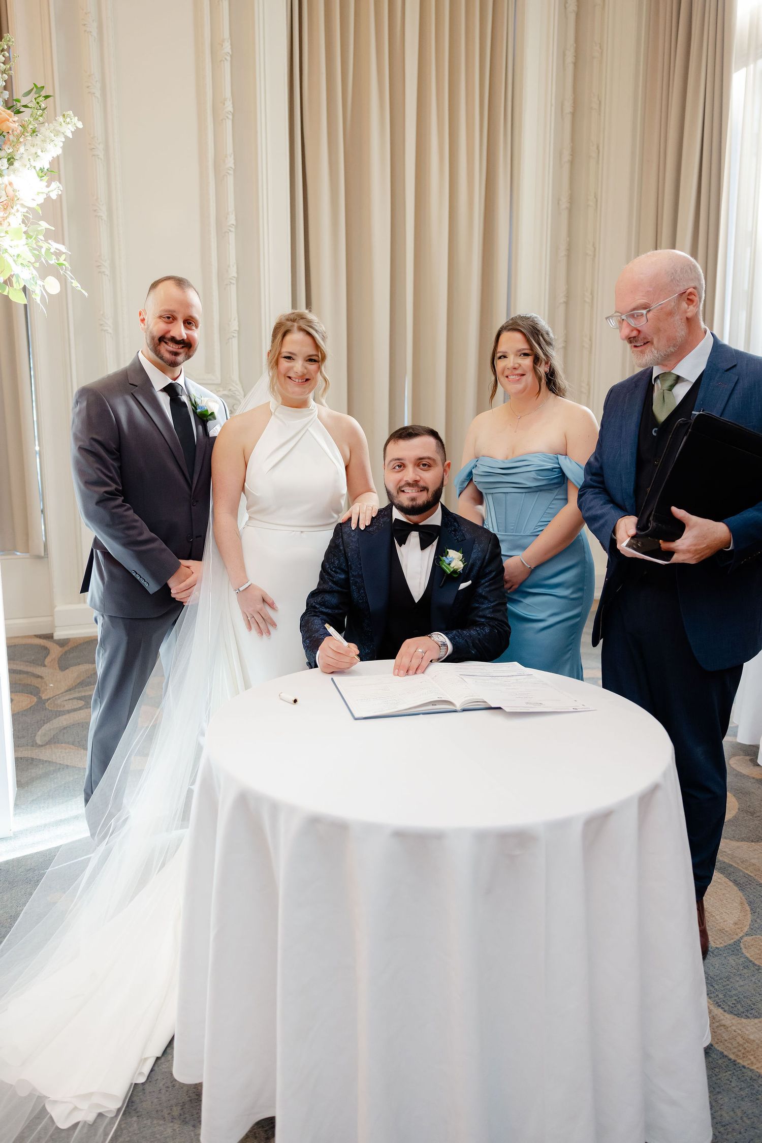 Wedding party gathered around white table for document signing in elegant ballroom setting.