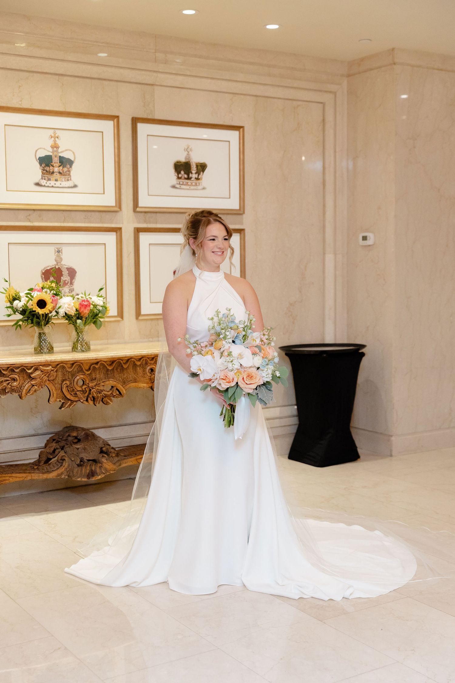 A bride in a flowing white wedding gown poses in an elegant room with framed artwork and floral arrangements.