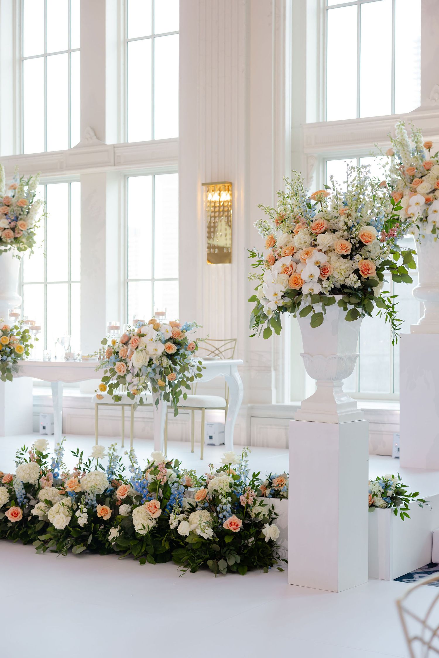 Luxurious wedding venue featuring peach and white floral arrangements beneath large windows.