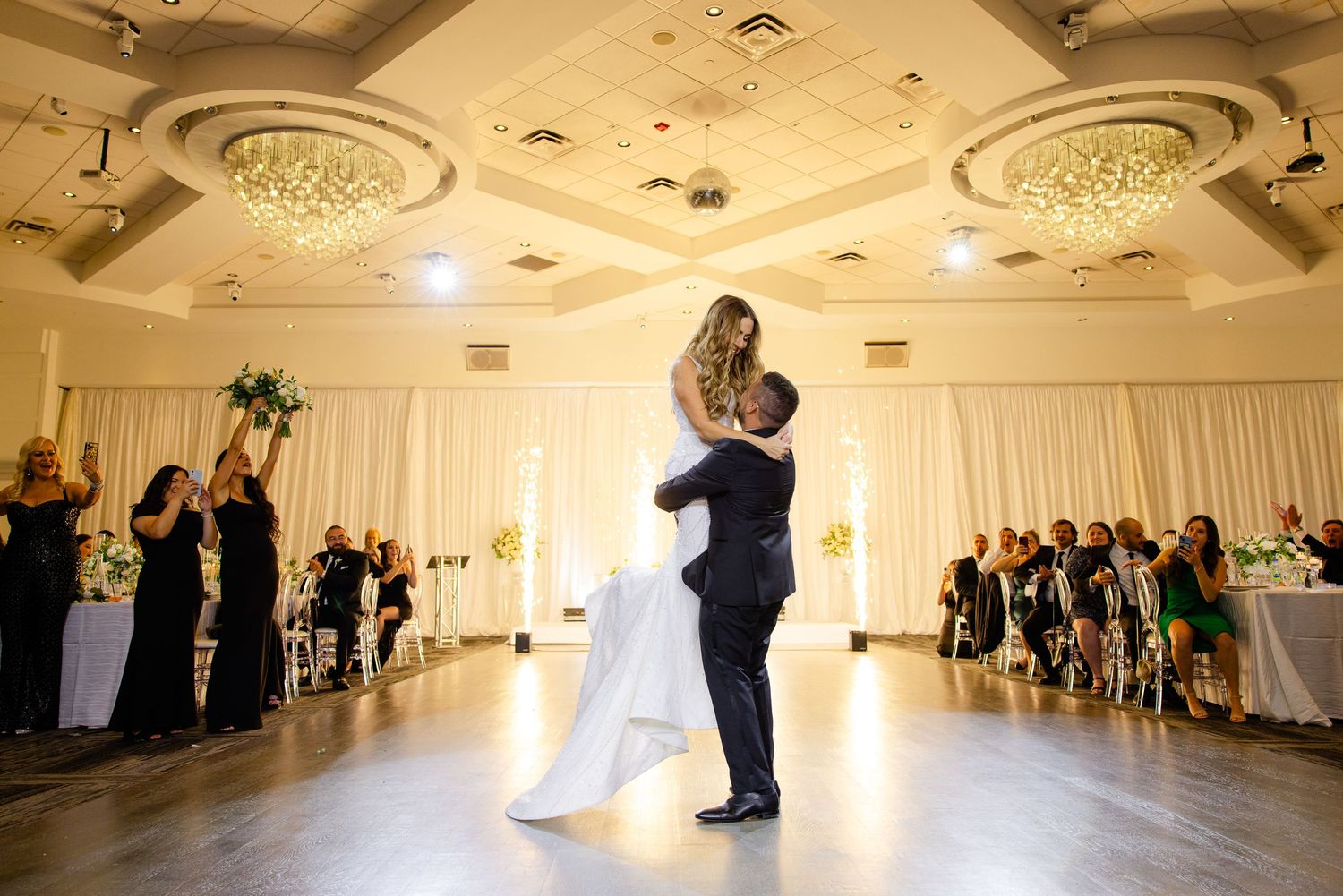 Romantic wedding dance moment captured in warm-lit ballroom with guests watching from tables.