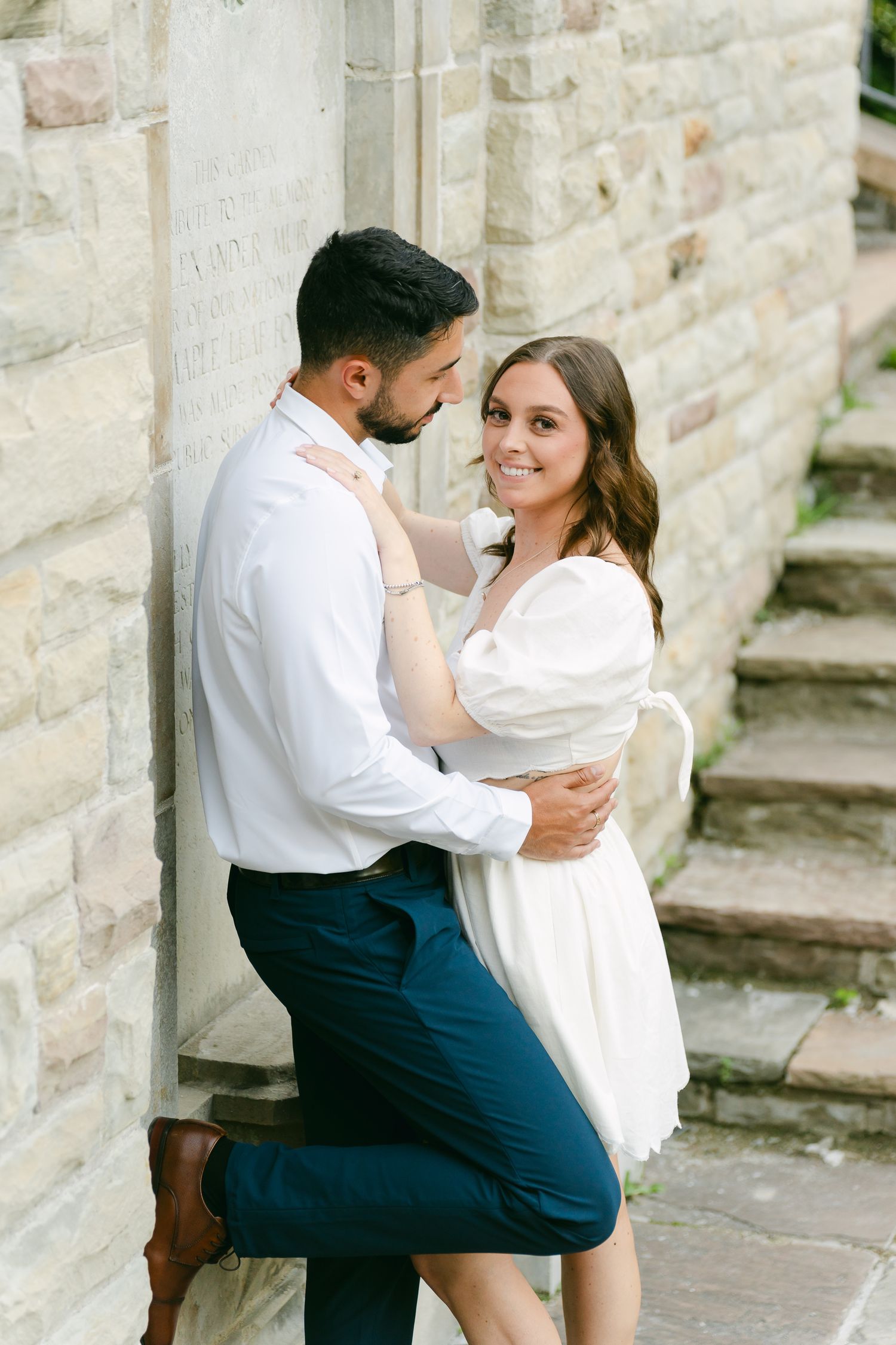 A romantic couple shares an intimate moment against a stone wall near historic steps.
