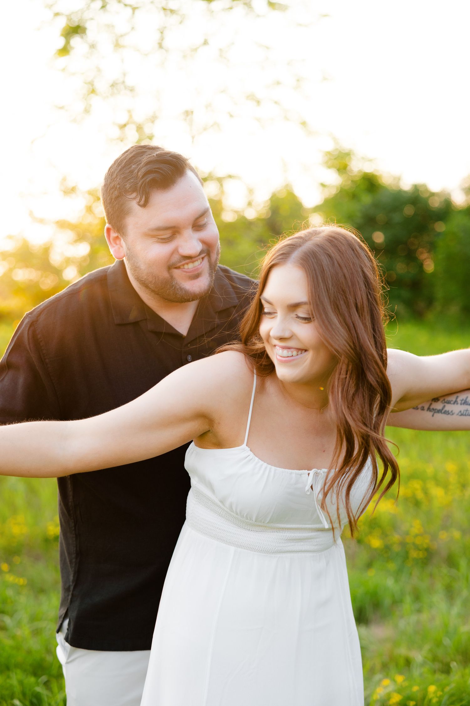 Playful couple poses in white summer dress and black shirt against golden sunset backdrop.