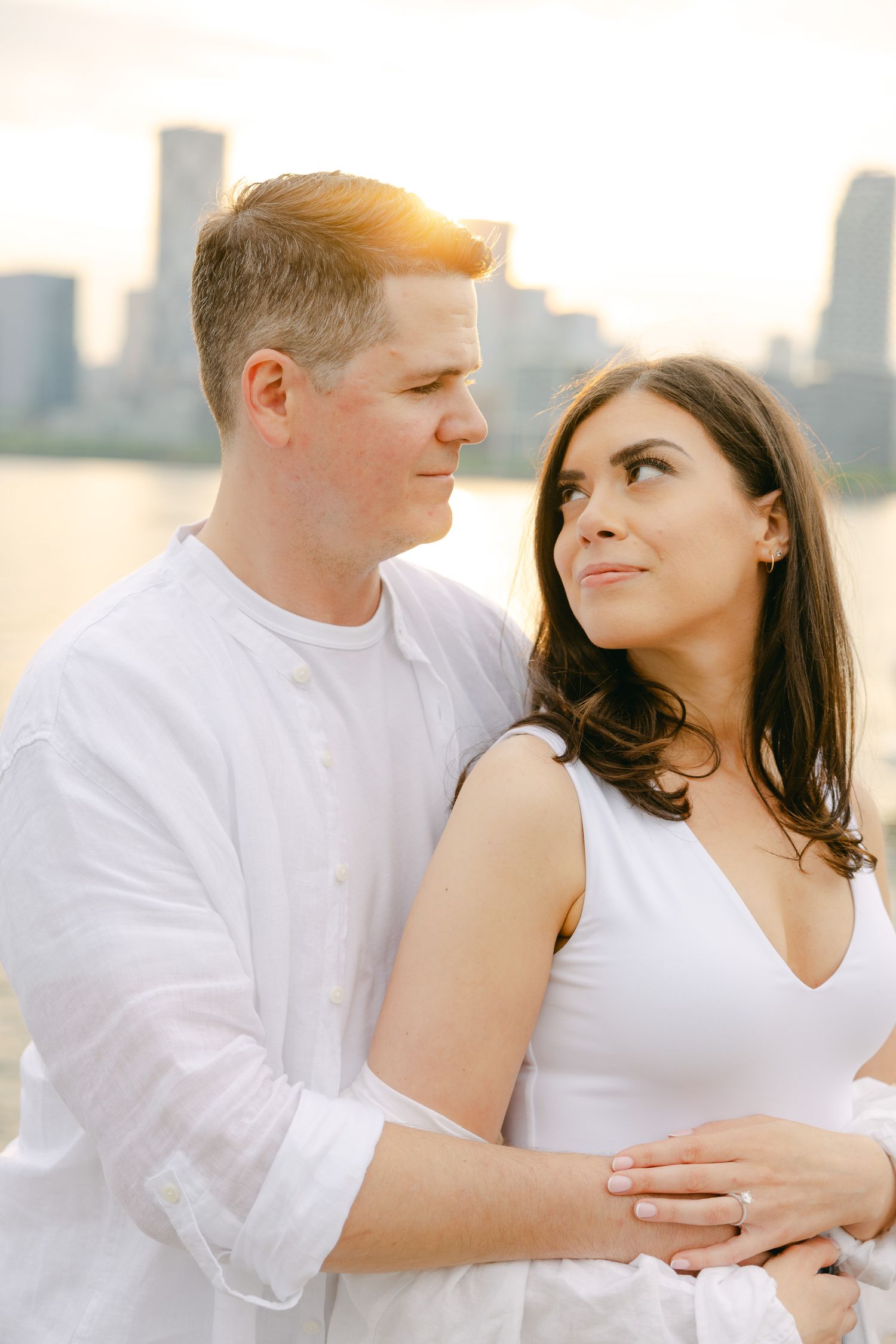 A sunset engagement photo with a couple in white clothing against a golden-lit city skyline.