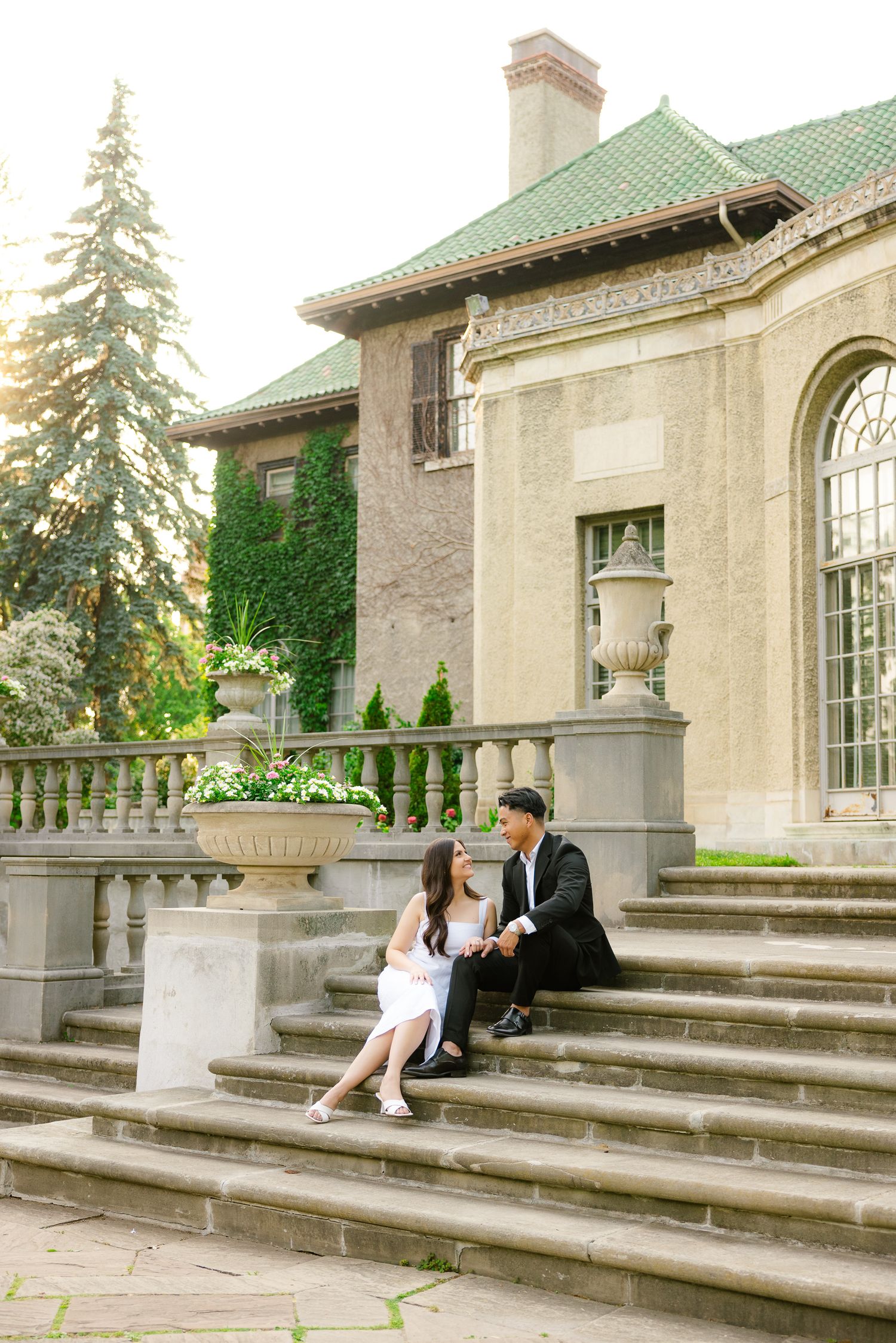 A couple shares an intimate moment on grand stone steps in front of a stately mansion with ornate architecture.