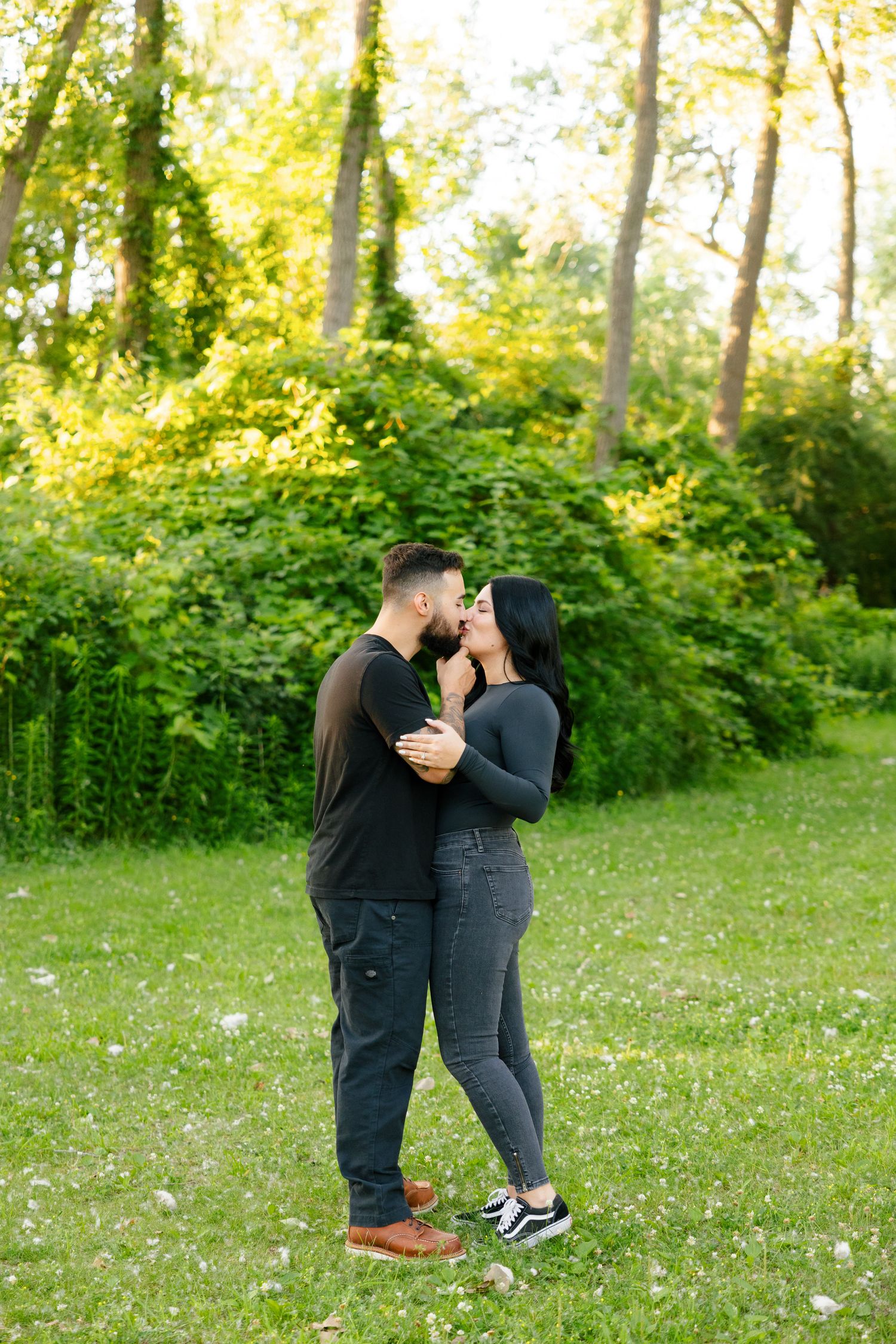 Couple embracing in a sunlit wooded area with lush green foliage in the background.