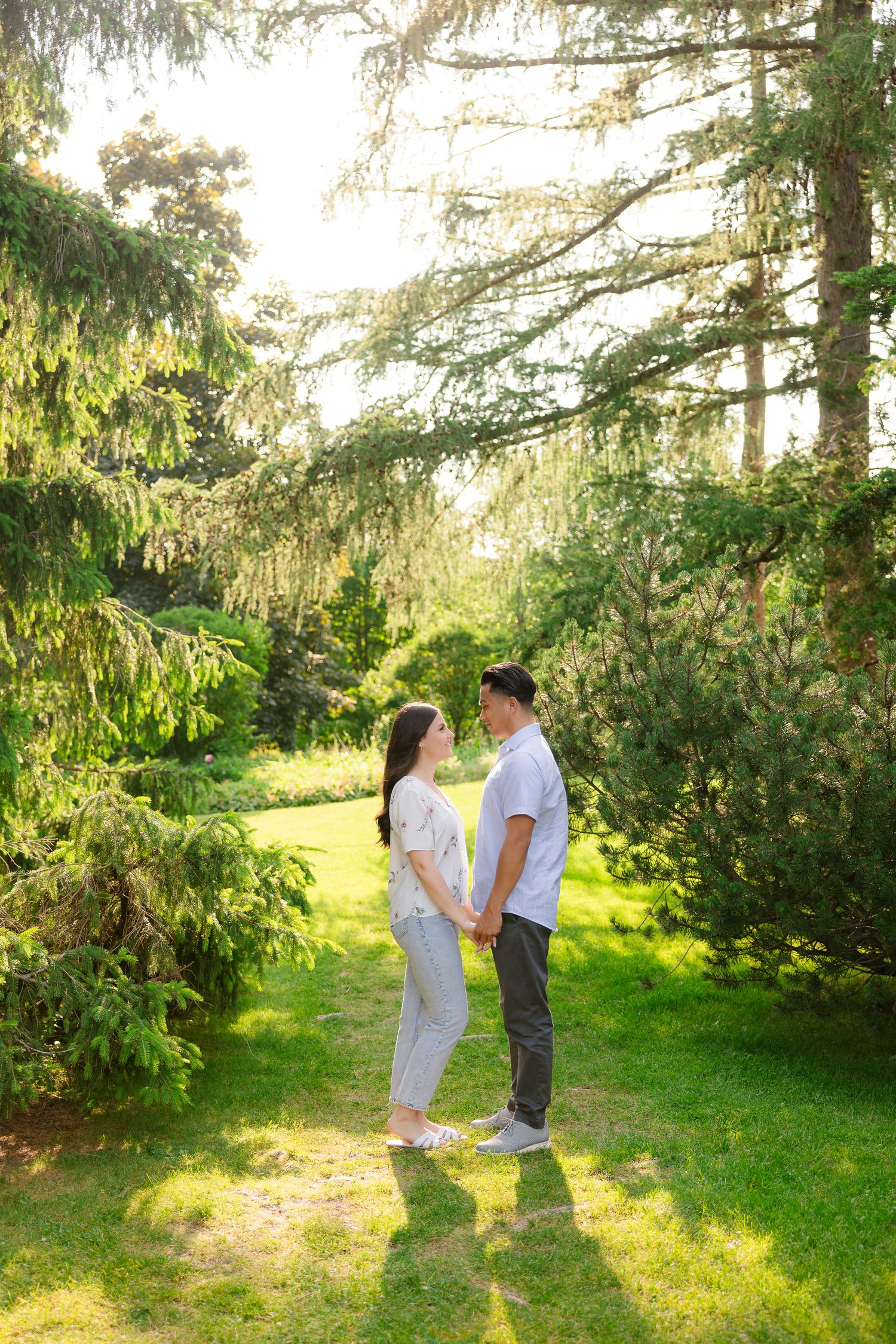 A romantic couple poses together in a dreamy garden setting with sunlight filtering through pine trees.