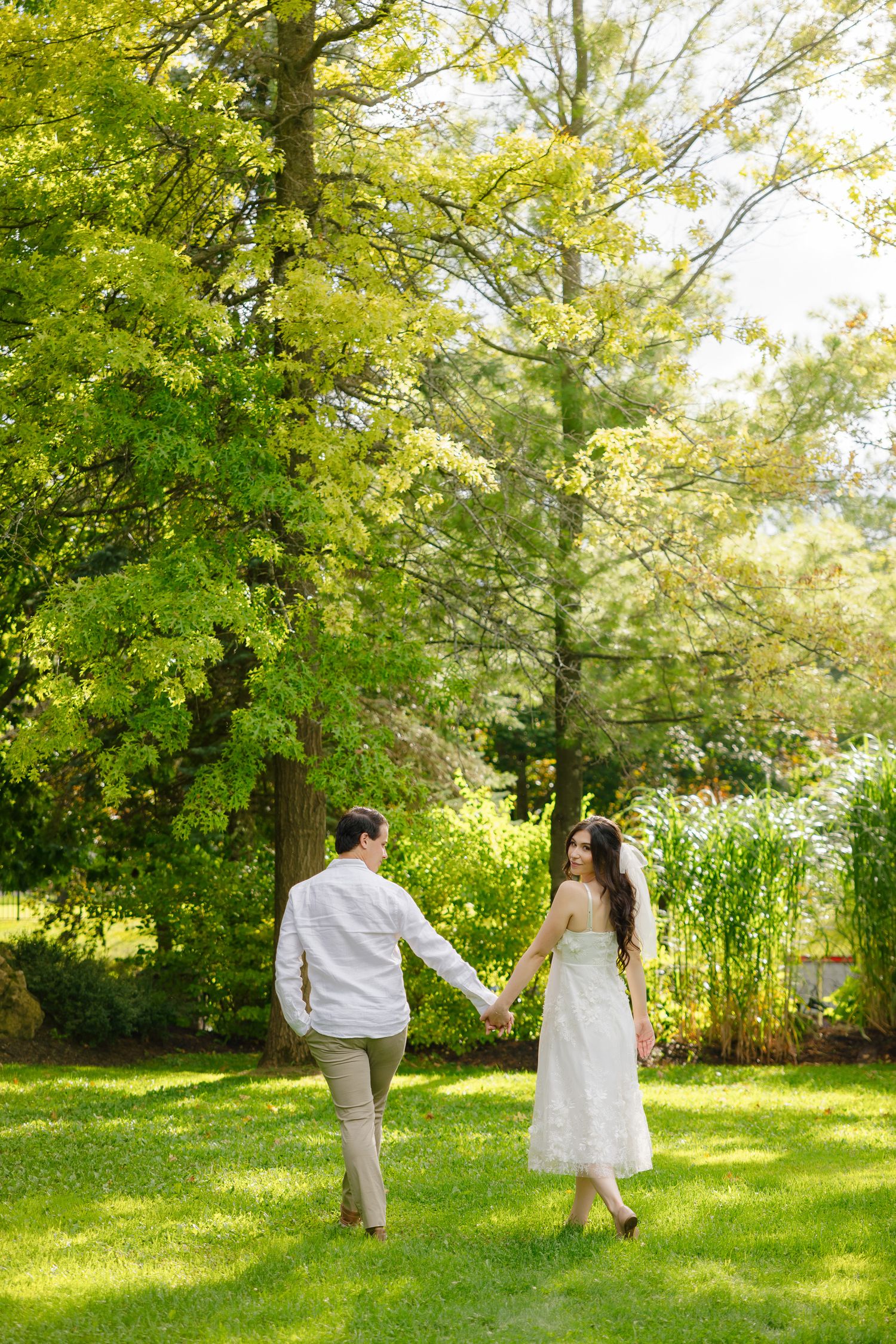 Wedding couple holding hands while walking through a sunny garden with flowering trees.
