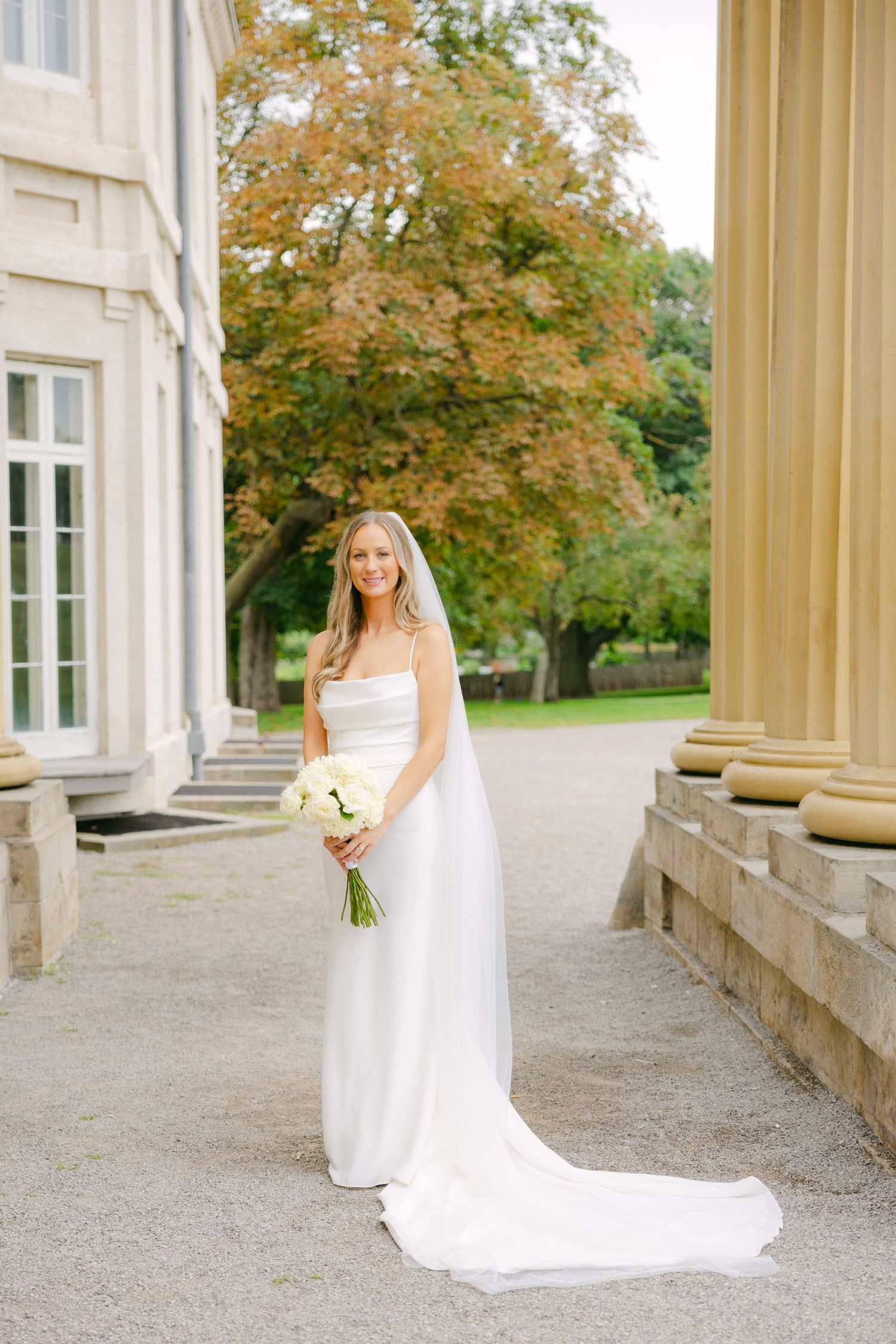 Elegant bride in white gown with long train poses between classic columns against autumn foliage backdrop.