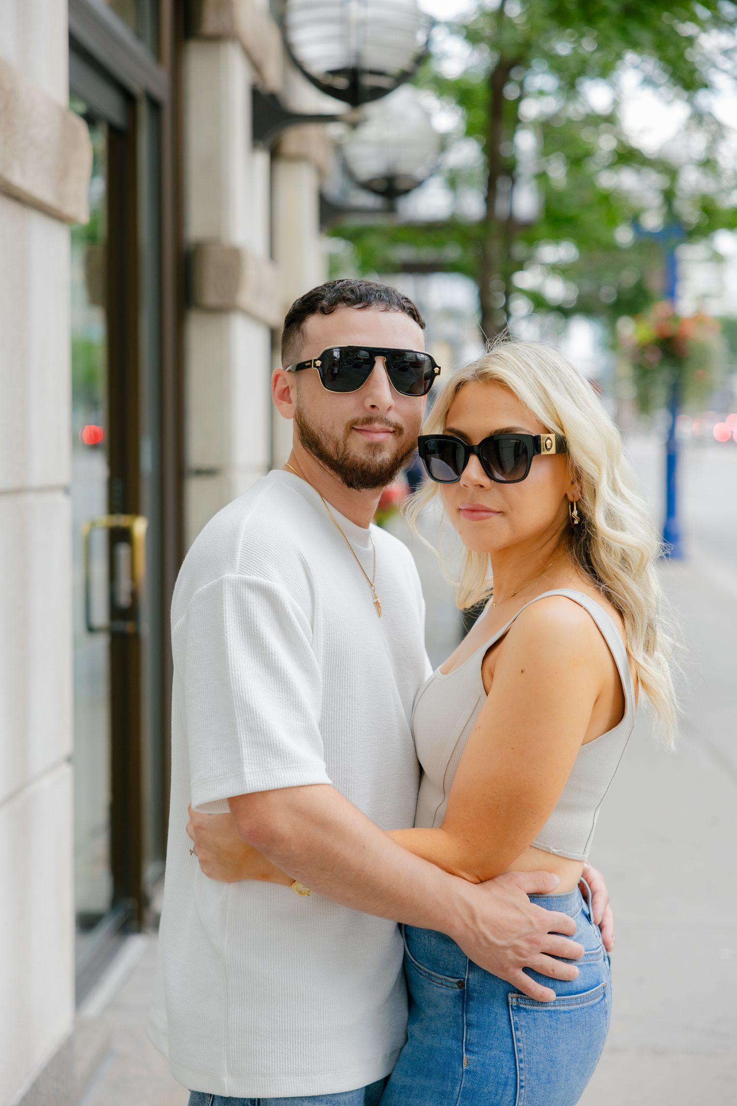 A couple in sunglasses embraces outside a building with classic street lamps.