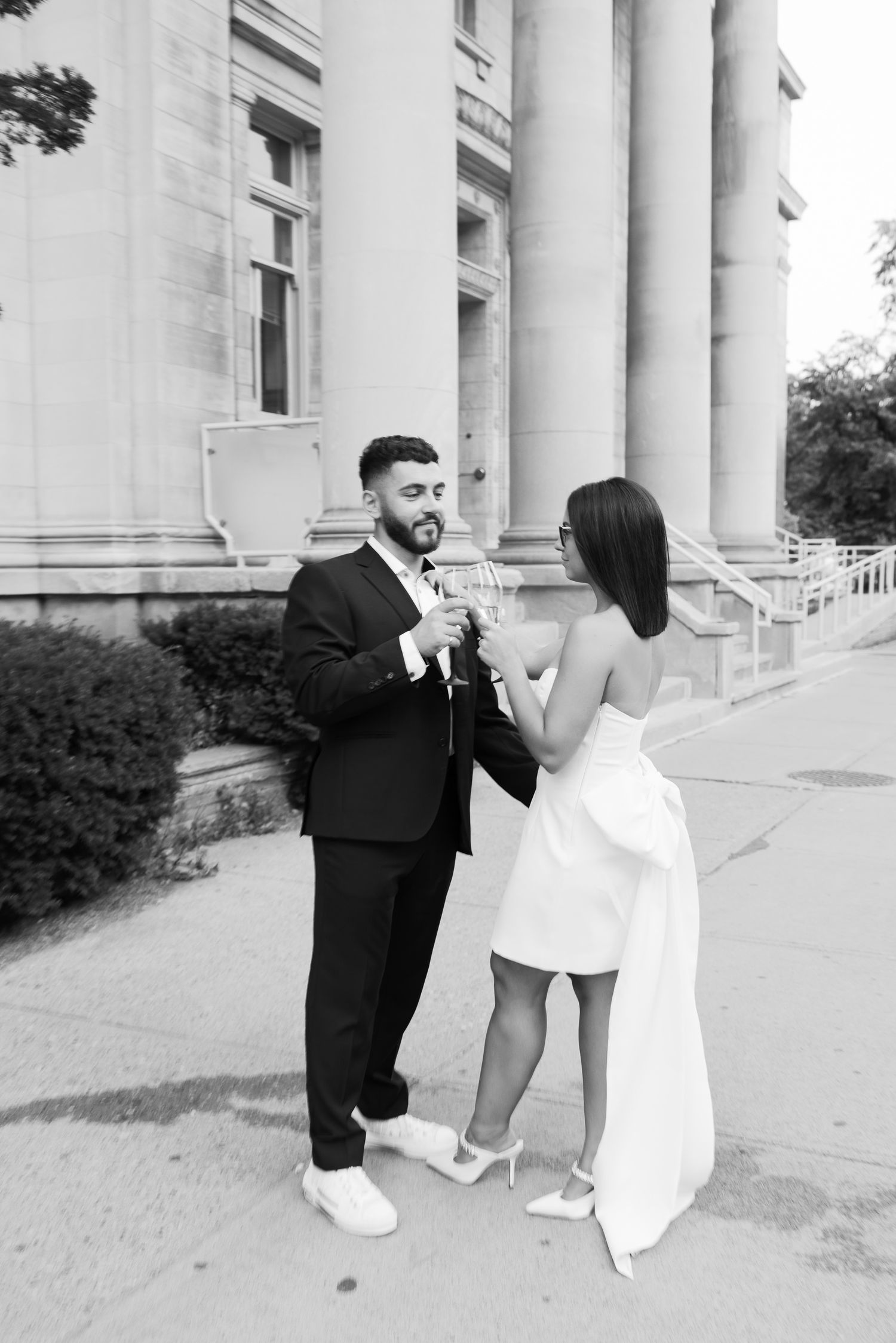 Couple in formal attire sharing drinks and laughter outside historic building with columns.