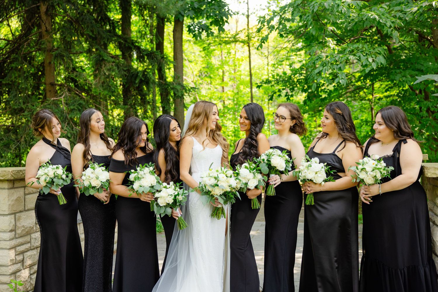 Wedding party smiling with white bouquets against stone wall and green foliage background.
