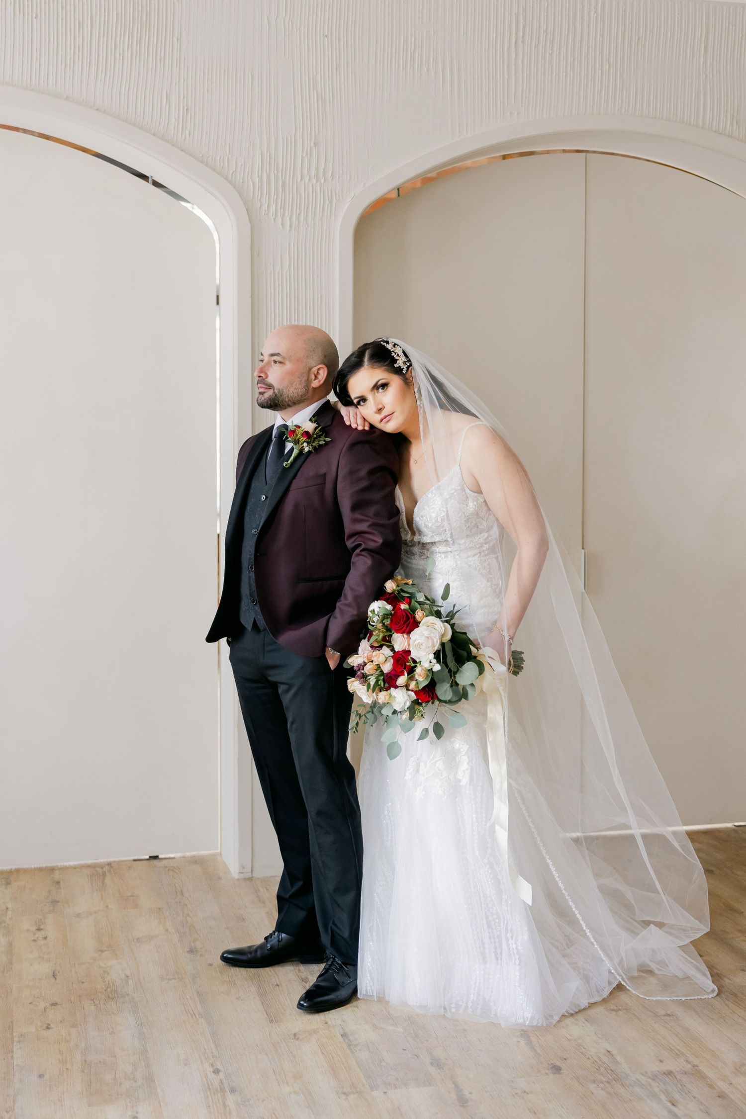 Stylish wedding couple poses against white wall with bride holding red and white bouquet.
