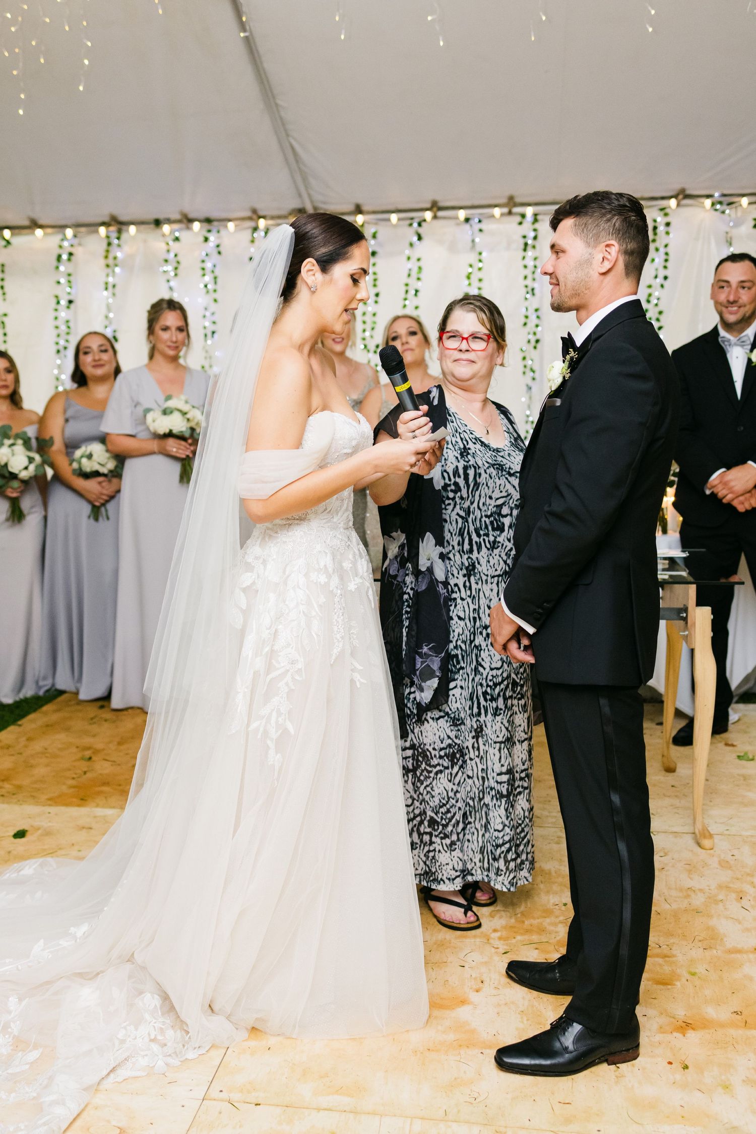 Wedding ceremony moment captured under tent with bridesmaids in grey dresses watching.