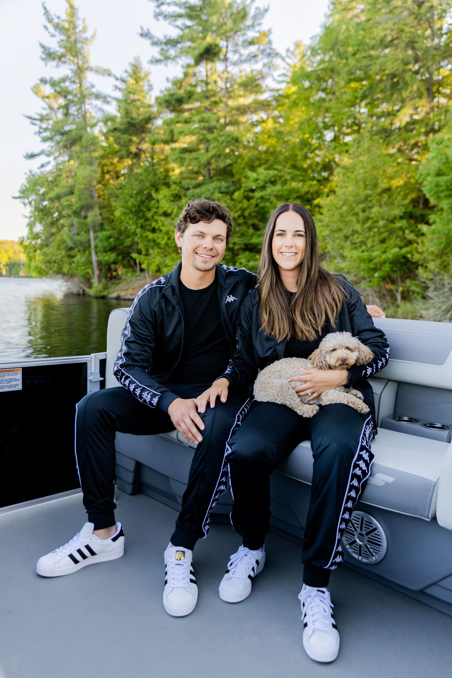 A couple in matching black Kappa tracksuits sit on a boat with their small dog against a scenic lake backdrop.