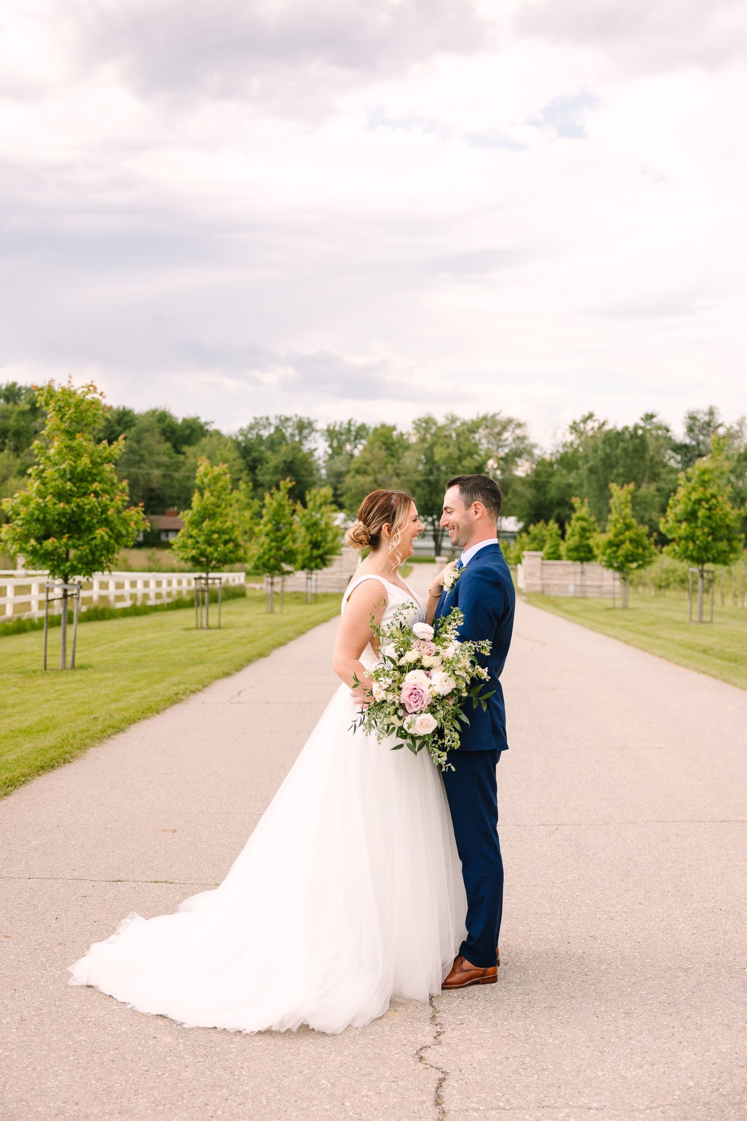 Wedding couple stands on path surrounded by green trees and white fencing.