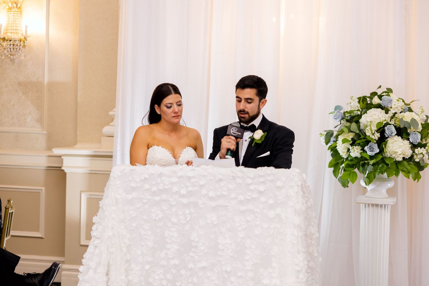 Newlyweds seated at ornate sweetheart table with white textured linens and floral arrangements.