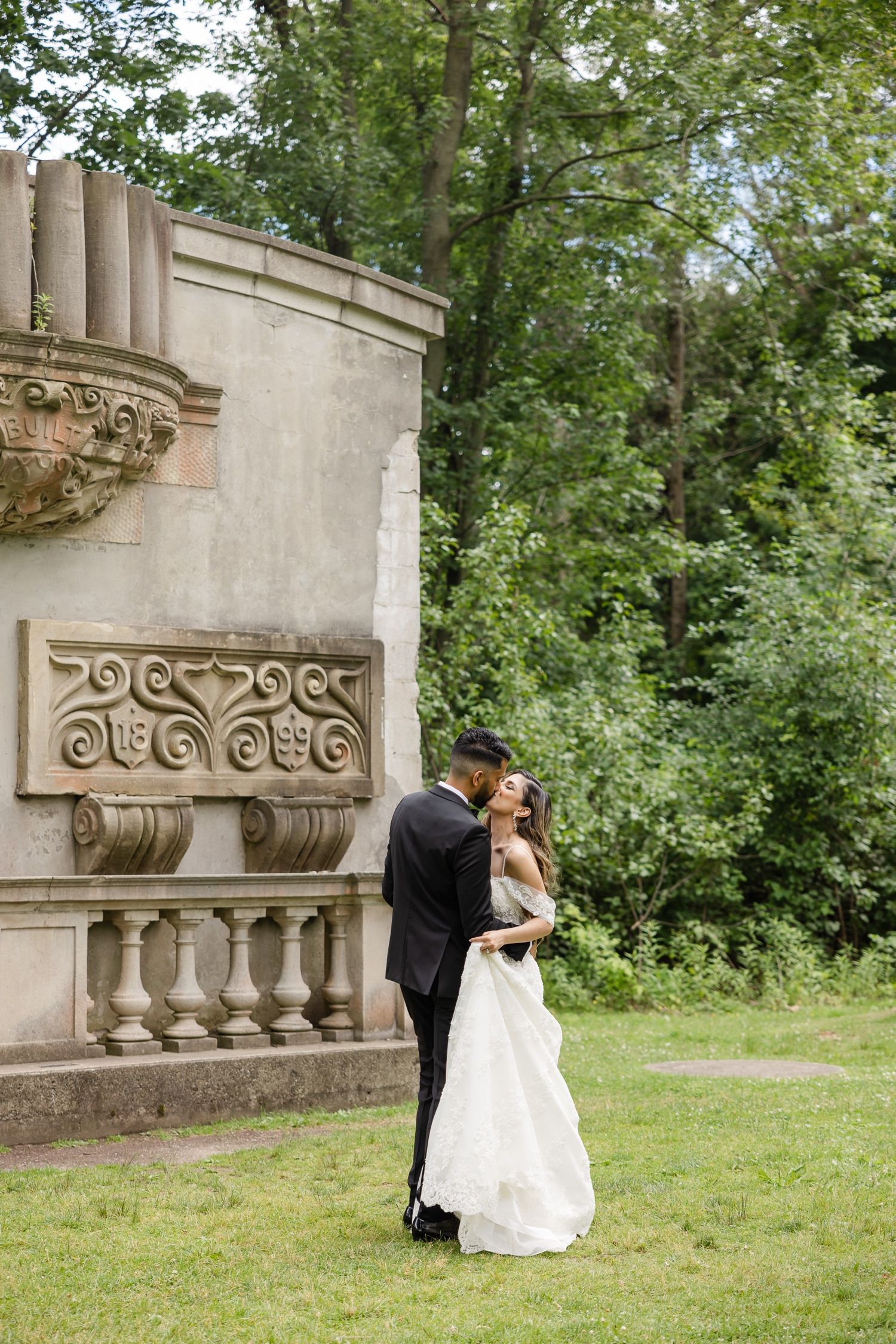 Elegant couple embraces near ornate stone architectural detail in lush garden setting.