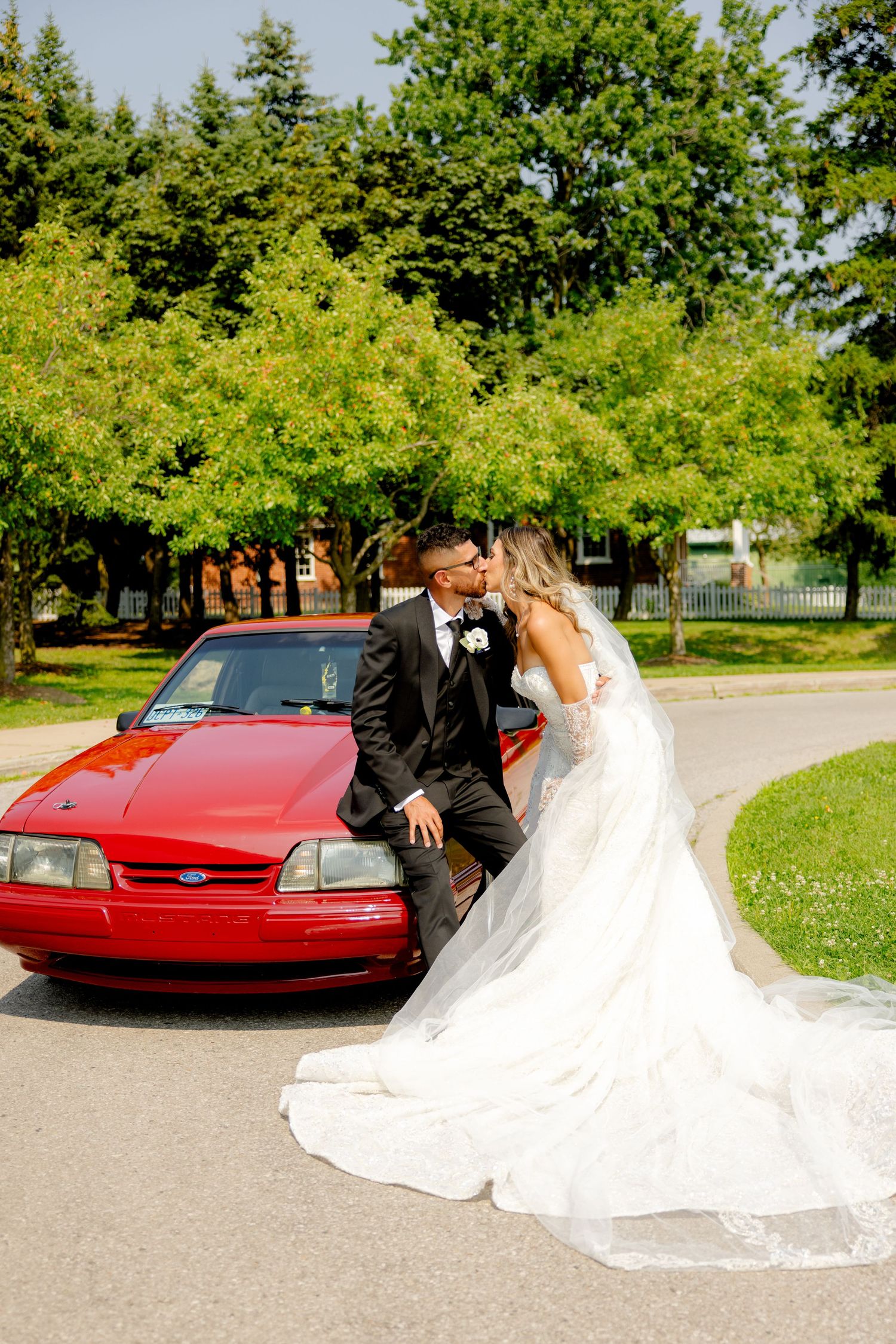 Sun-lit wedding portrait featuring a red car and beautiful garden scenery.