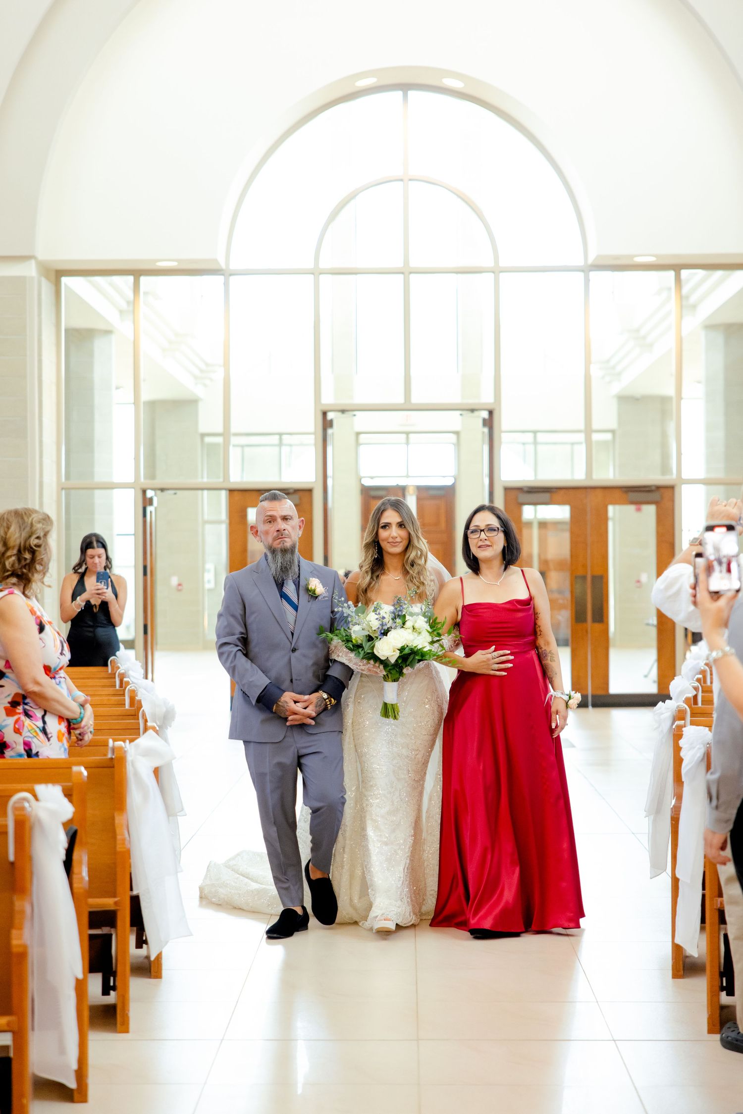 A wedding processional in a sunlit church with elegant arched windows and wooden pews.